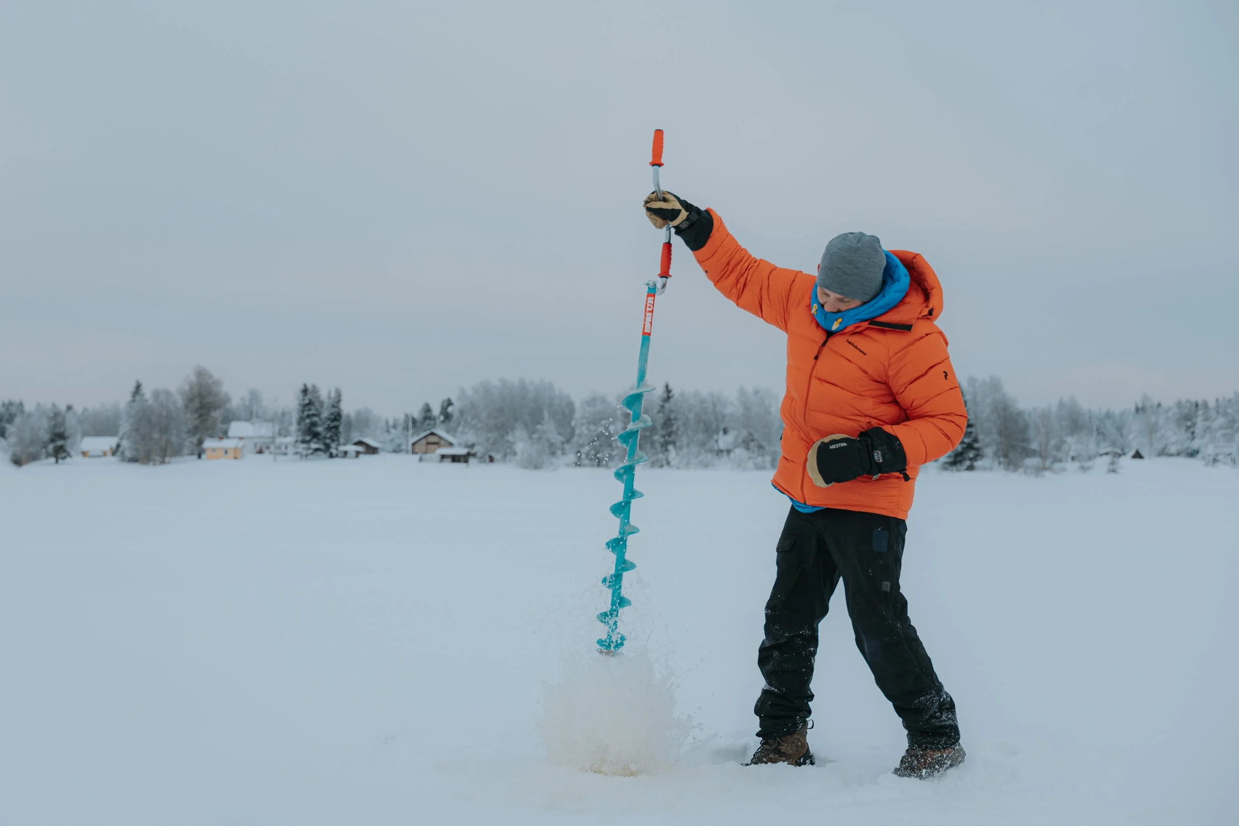 Person in orange winter jacket and gray beanie using a handheld ice auger in a snowy landscape with trees and small houses in the background.