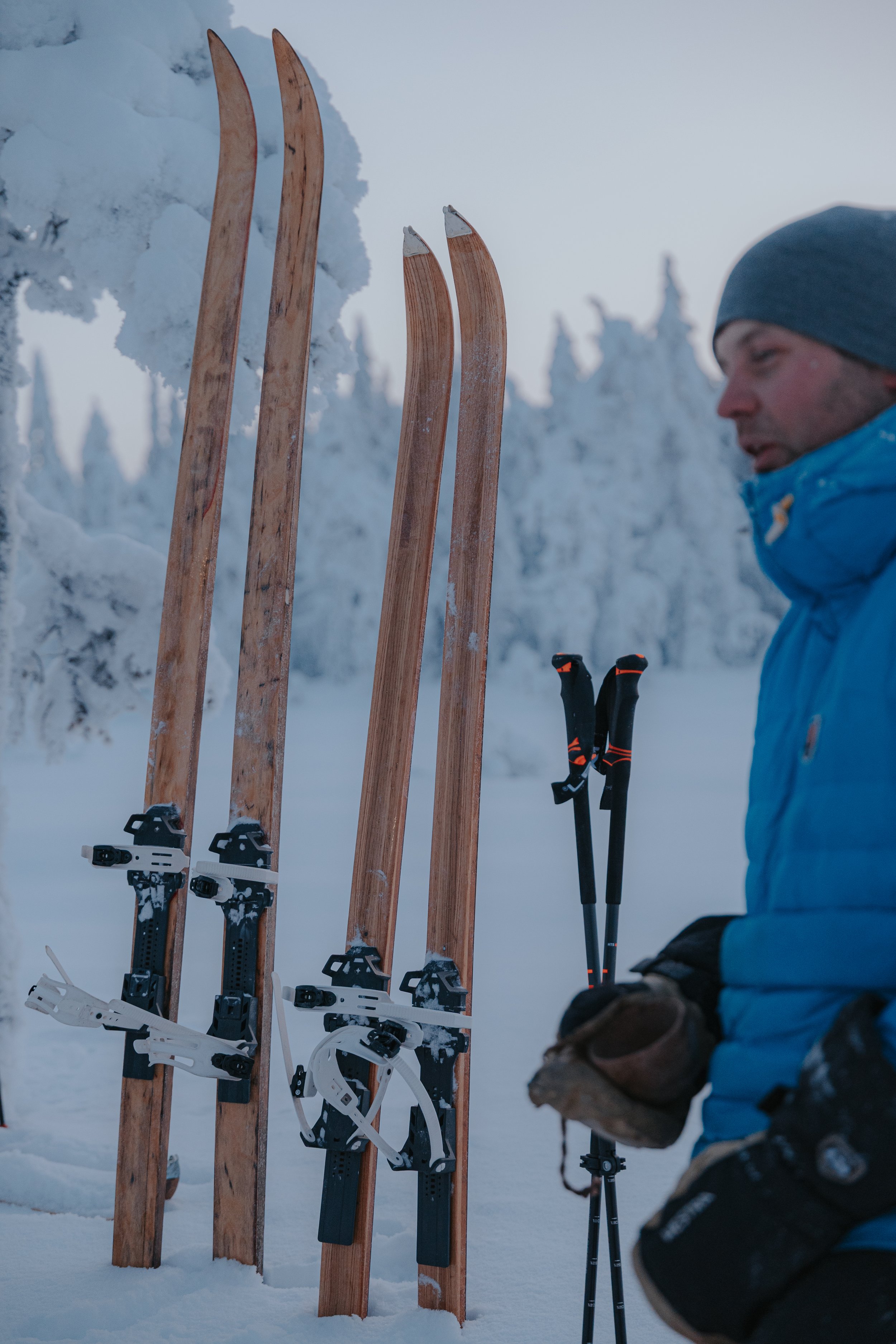 Close-up of four wooden skis with ski bindings, and a person holding ski poles in a snowy forest landscape during winter.