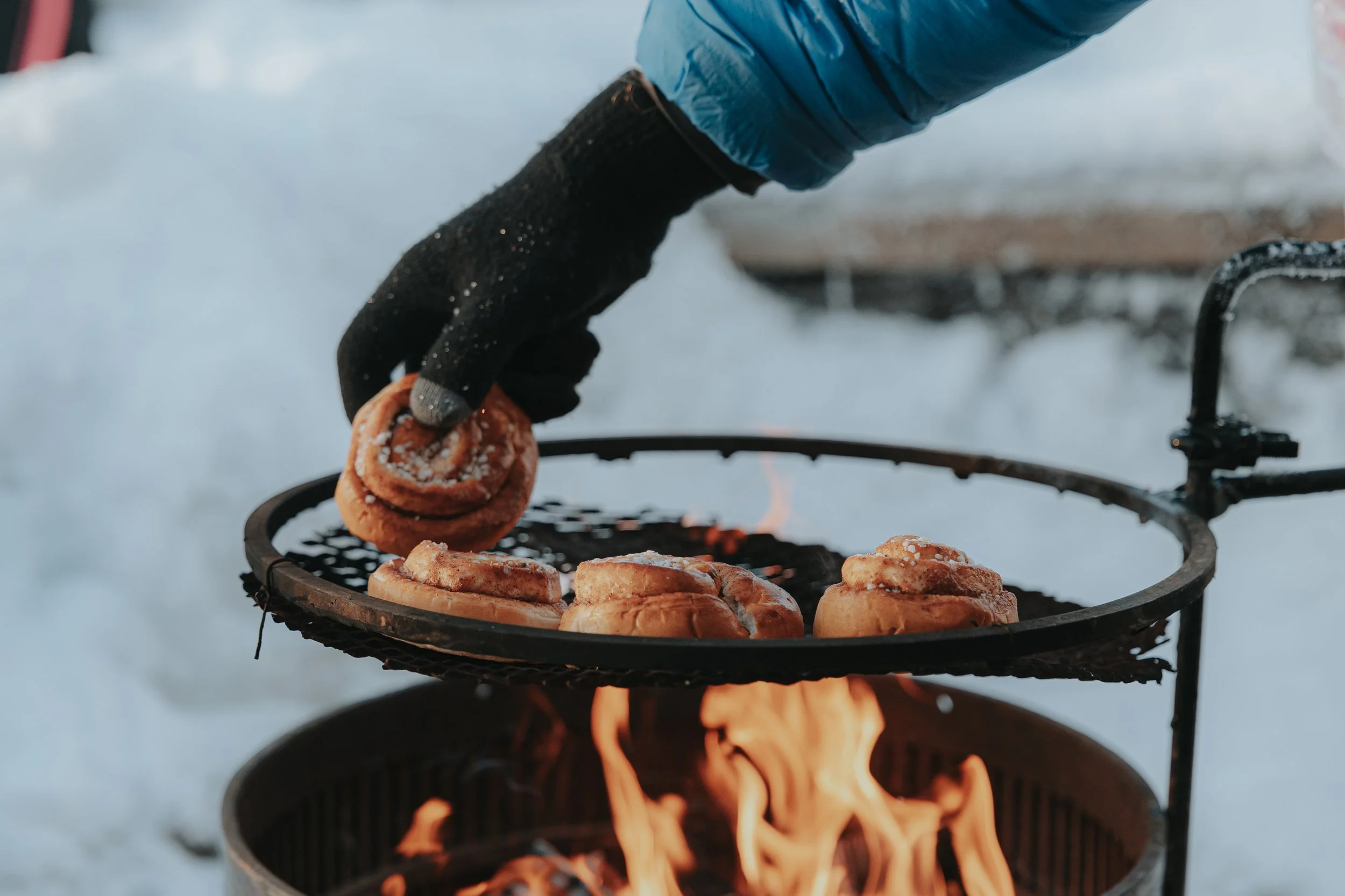 Person wearing black gloves and a blue jacket placing a cinnamon roll on a grill basket above a campfire outdoors, with snow visible in the background.