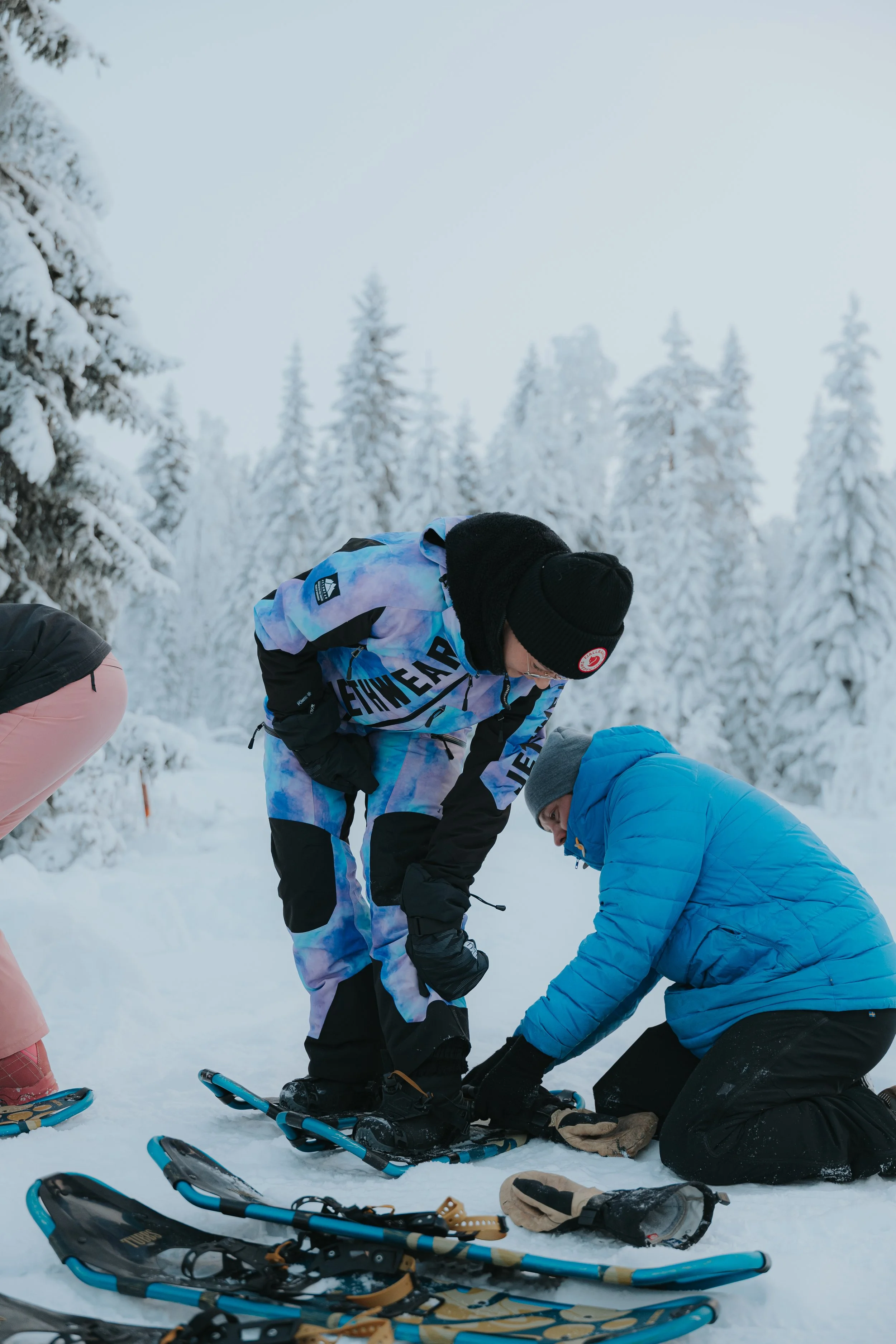 Group of people in winter clothing preparing snowshoes outdoors in a snowy forest.