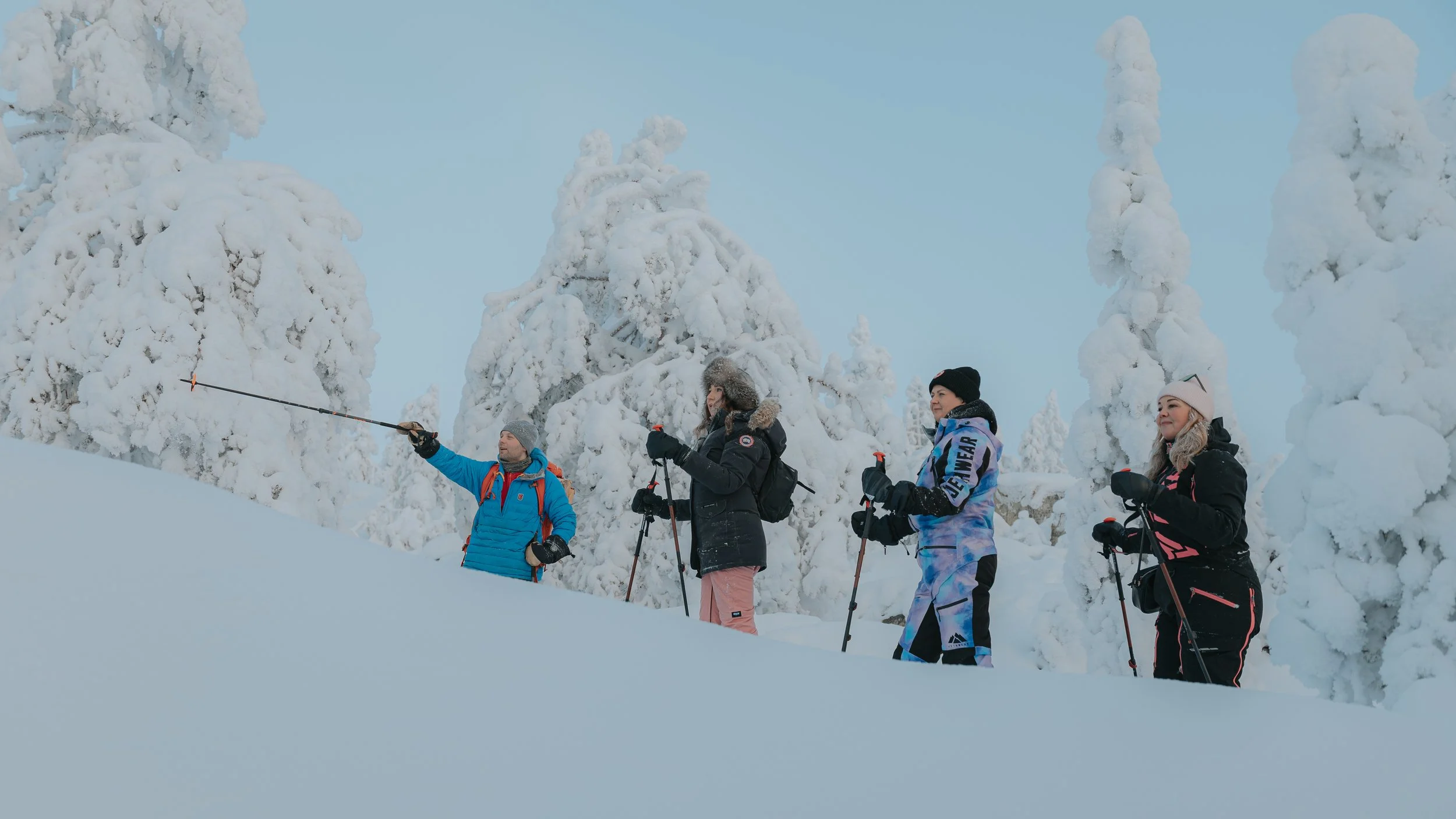 Four people in winter clothing standing in snowy forest with snow-covered trees, some holding ski poles, one person pointing in the distance.