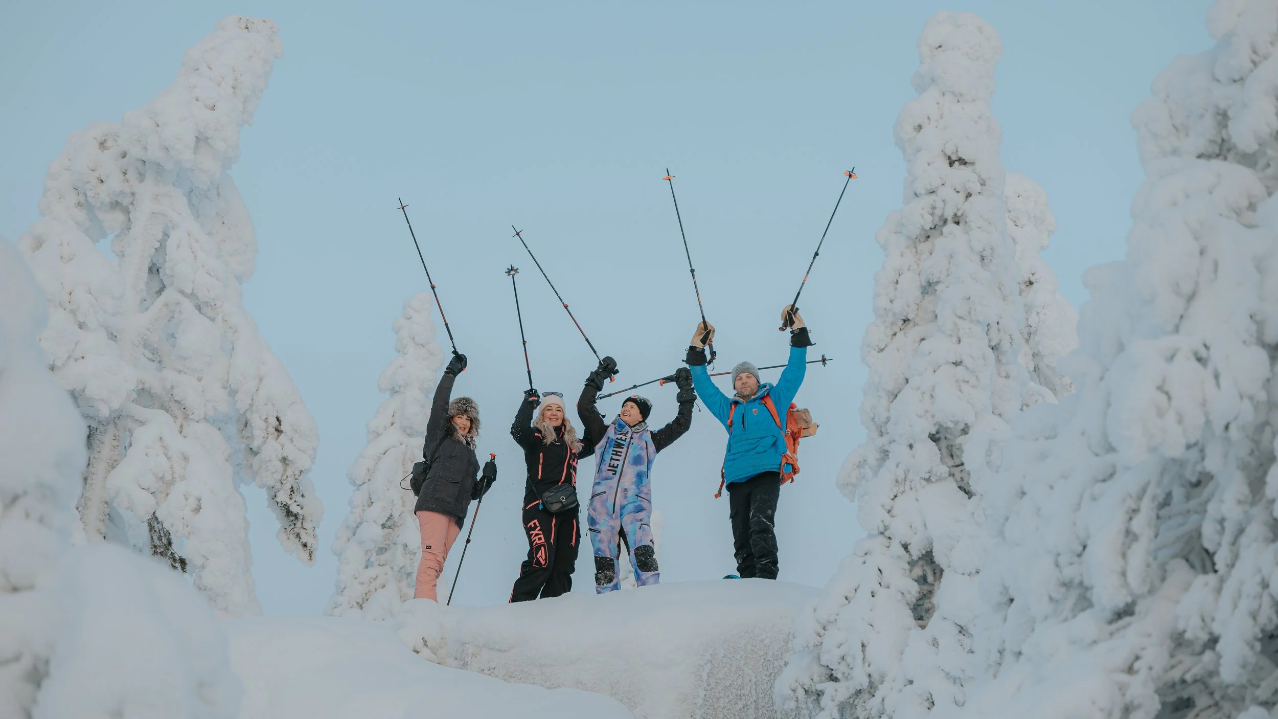 Four people in winter attire standing amidst snow-covered trees, holding ski poles up in celebration outdoors.