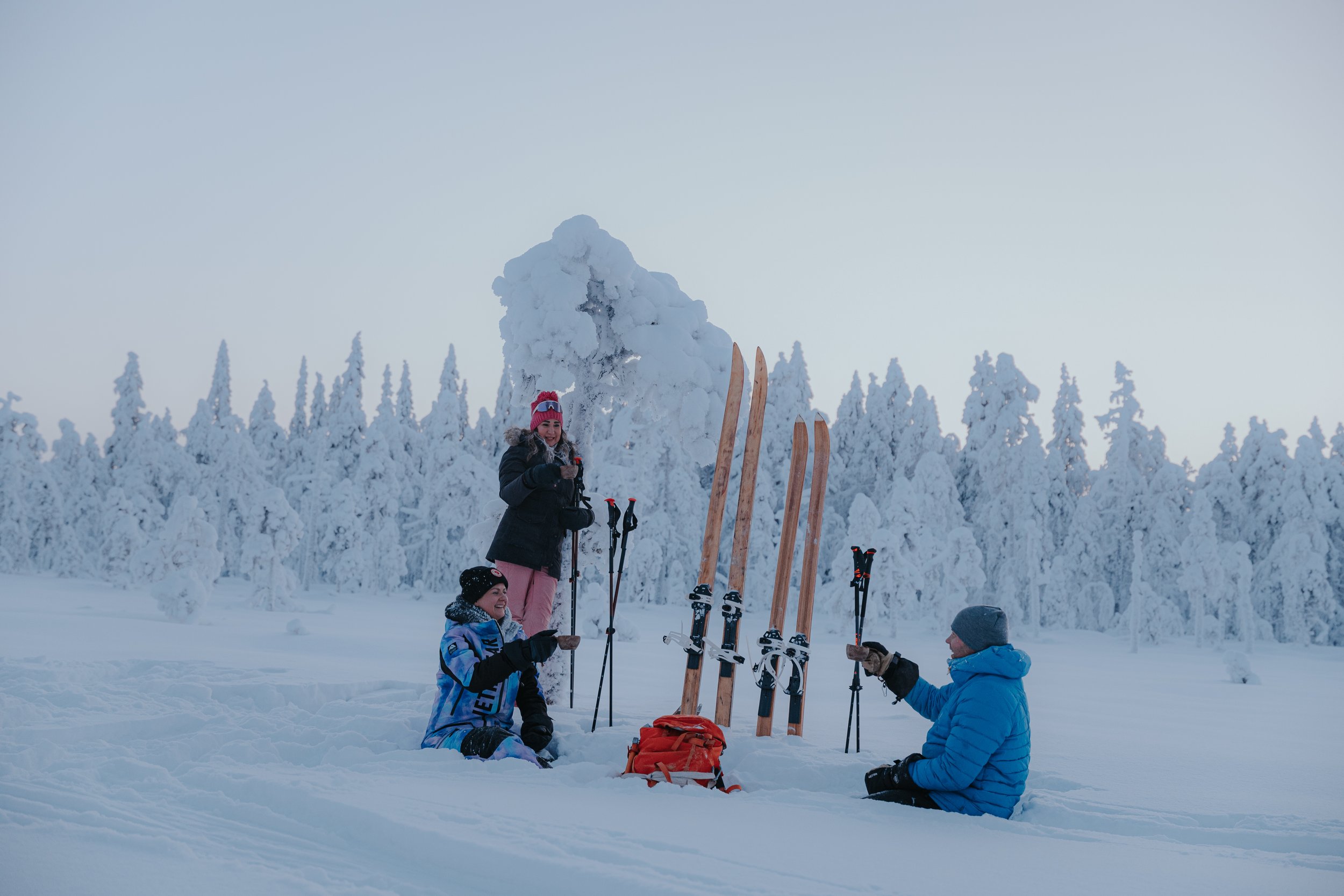 People in winter clothing sitting in the snow with skis upright in the background, surrounded by snow-covered trees under a pale sky.