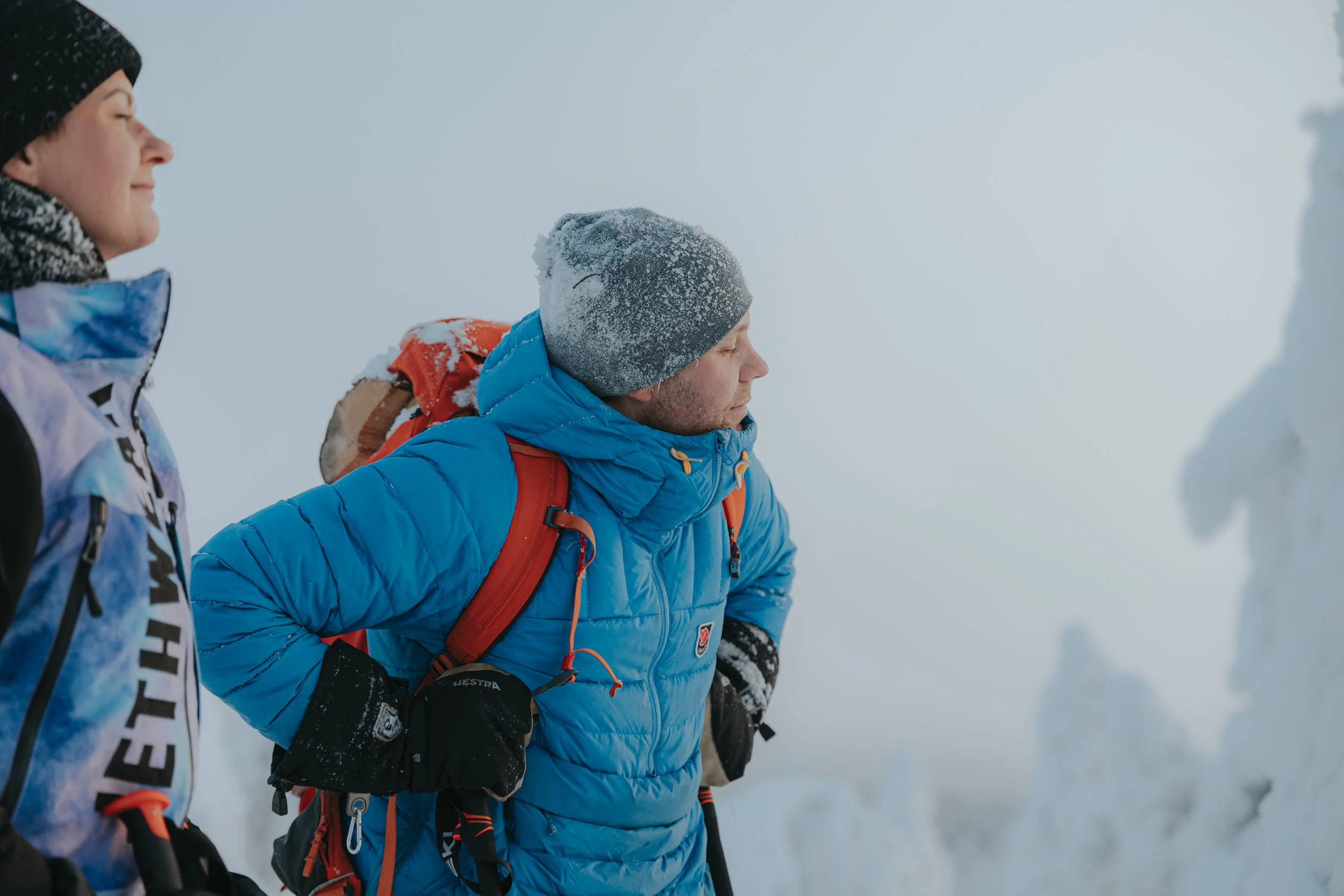 Two people dressed in winter outdoor gear, standing in a snowy landscape with fog and snow-covered rocks or ice in the background.