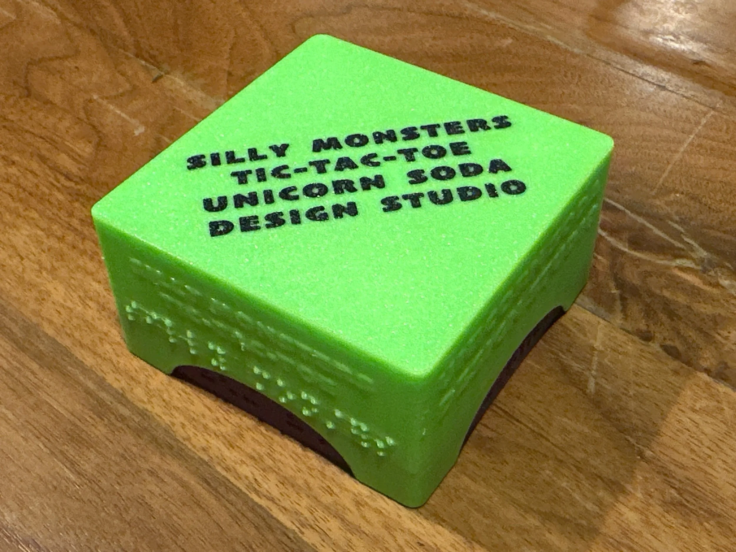 Closed box on a wooden surface. Box top is neon green and the base is purple. Braille and print labeling on the sides, and print only on the top surface of the box.