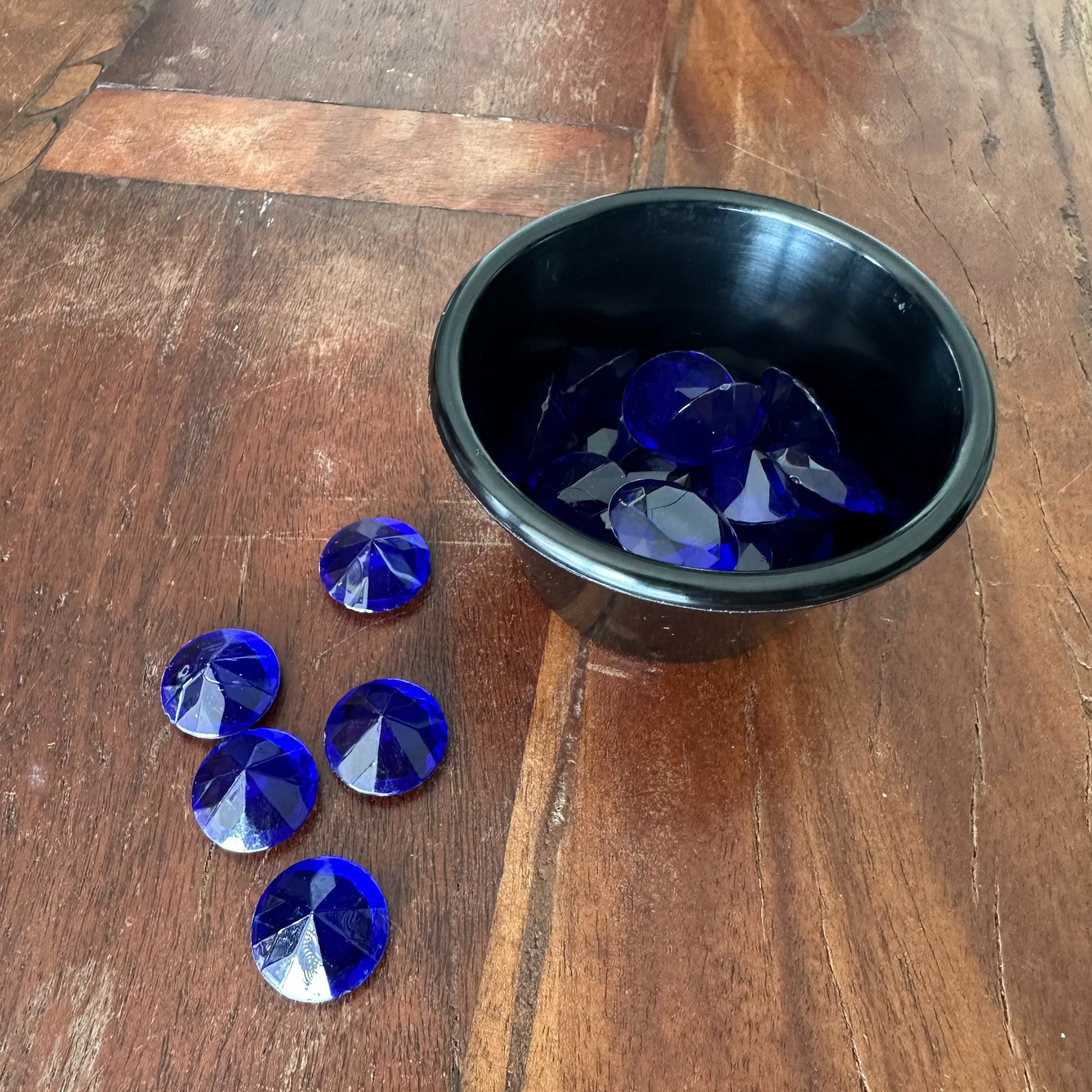 Sapphire blue acrylic gems shown in and scattered around a circular cup, against a dark wood table.