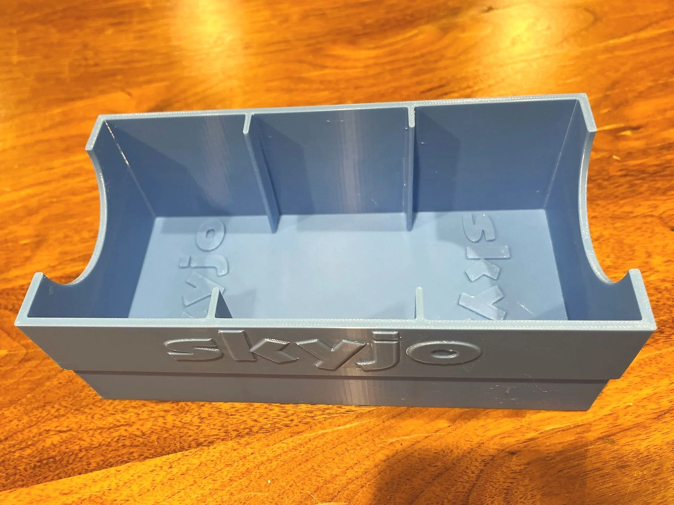 Blue card box insert, with SKYJO in print facing out. The insert has 3 sections for cards, and a semi-circular cut out at each of the ends.