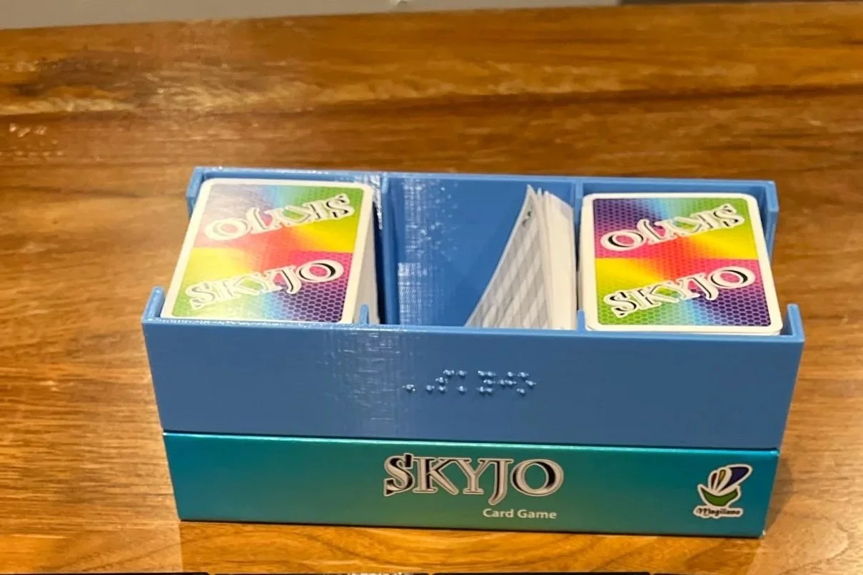 SKYJO insert with braille label facing outward. Insert is shown inside manufacturer's box, with cards in the 2 end compartments. The center compartment is empty.