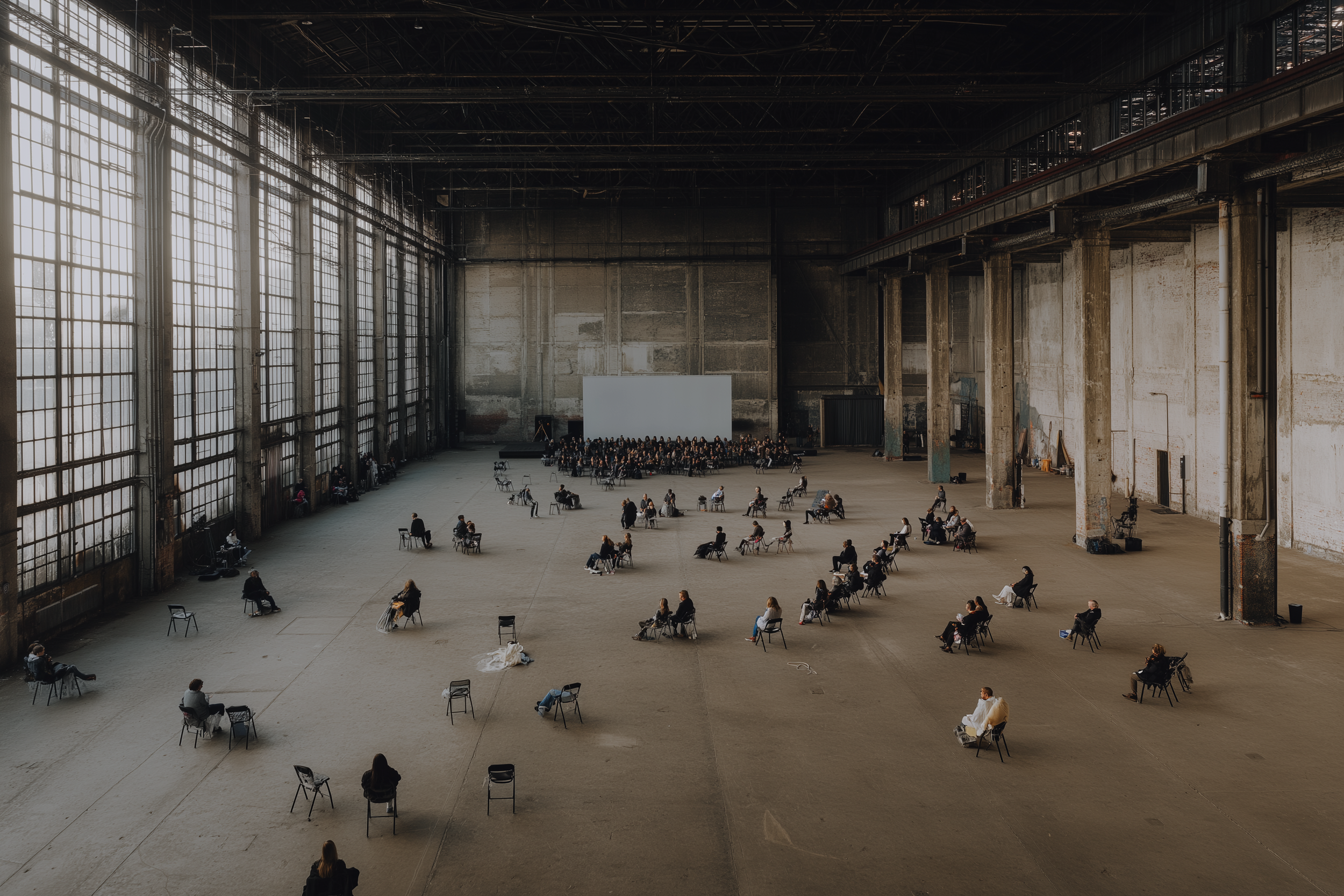 Une grande salle industrielle avec de hauts plafonds et de grandes fenêtres en verre. Des personnes sont assises seules ou en petits groupes, espacées les unes des autres, sur des chaises disposées dans toute la salle, donnant l'impression d'une séan