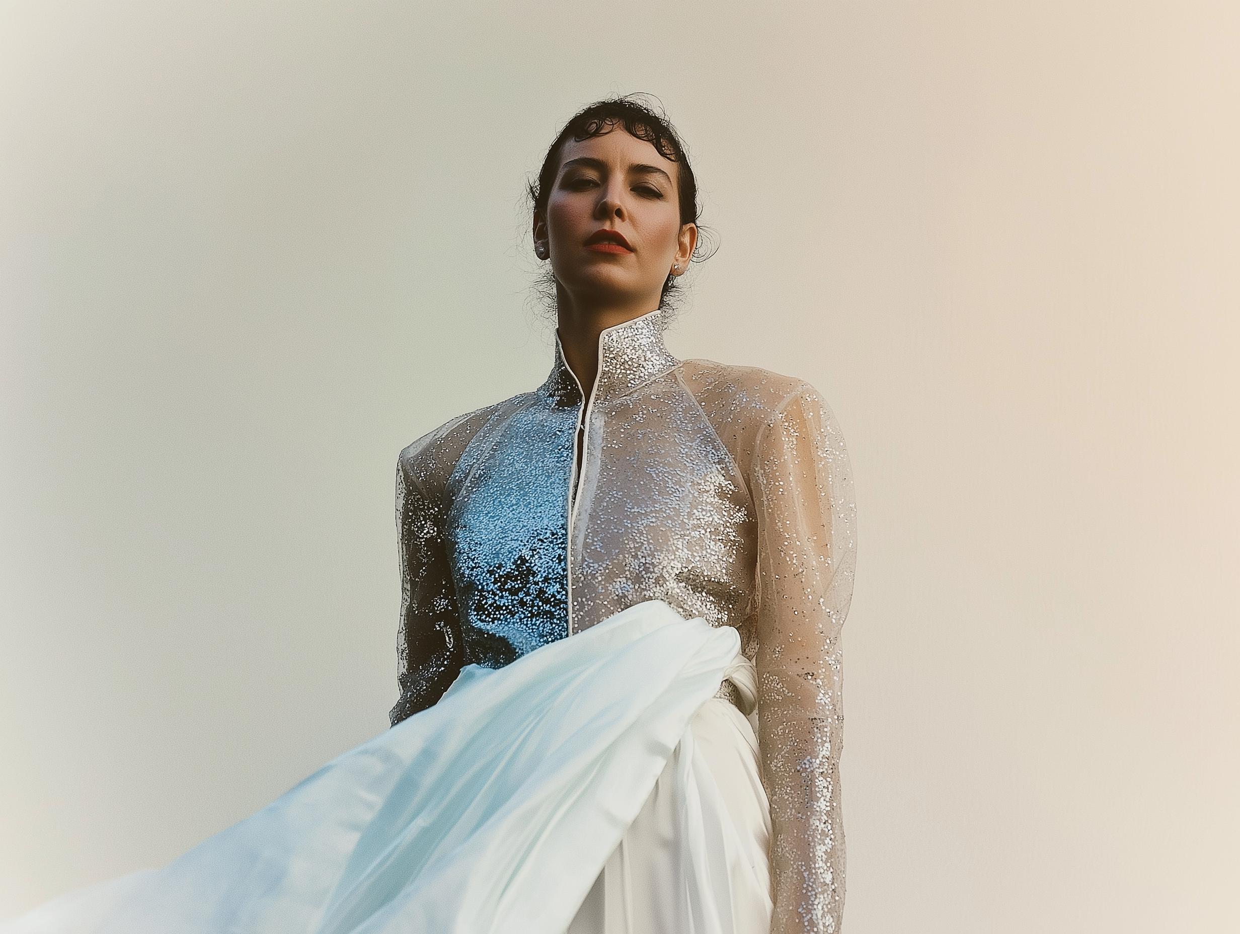 Femme portant une robe élégante avec une veste transparente en sequins, tenant un tissu blanc avant un fond clair.