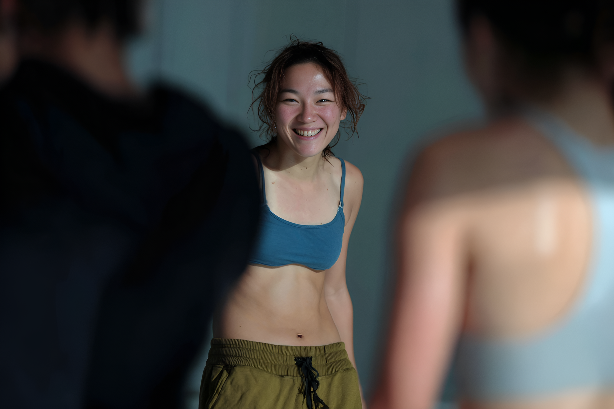 Une jeune femme souriante en tenue décontractée, portant un top bleu et un pantalon kaki, entourée de deux autres personnes floues, dans un environnement intérieur.