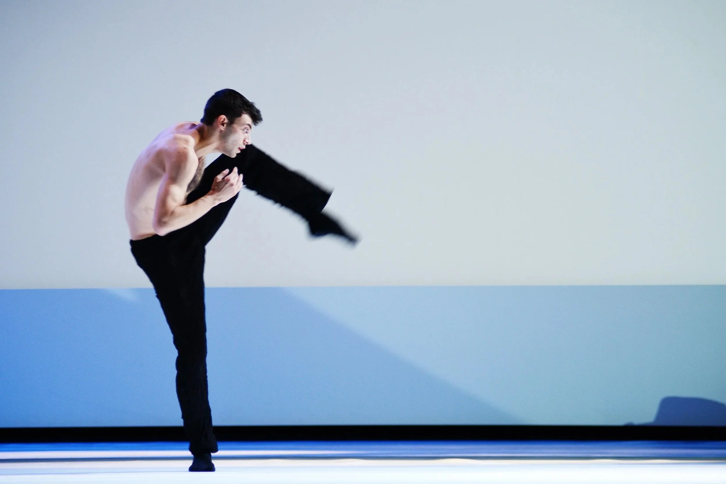 Un danseur en tailleur noir effectuant un saut acrobatique avec un pied en l'air et un bras plié, dans un espace minimaliste avec un fond blanc et un sol gris.