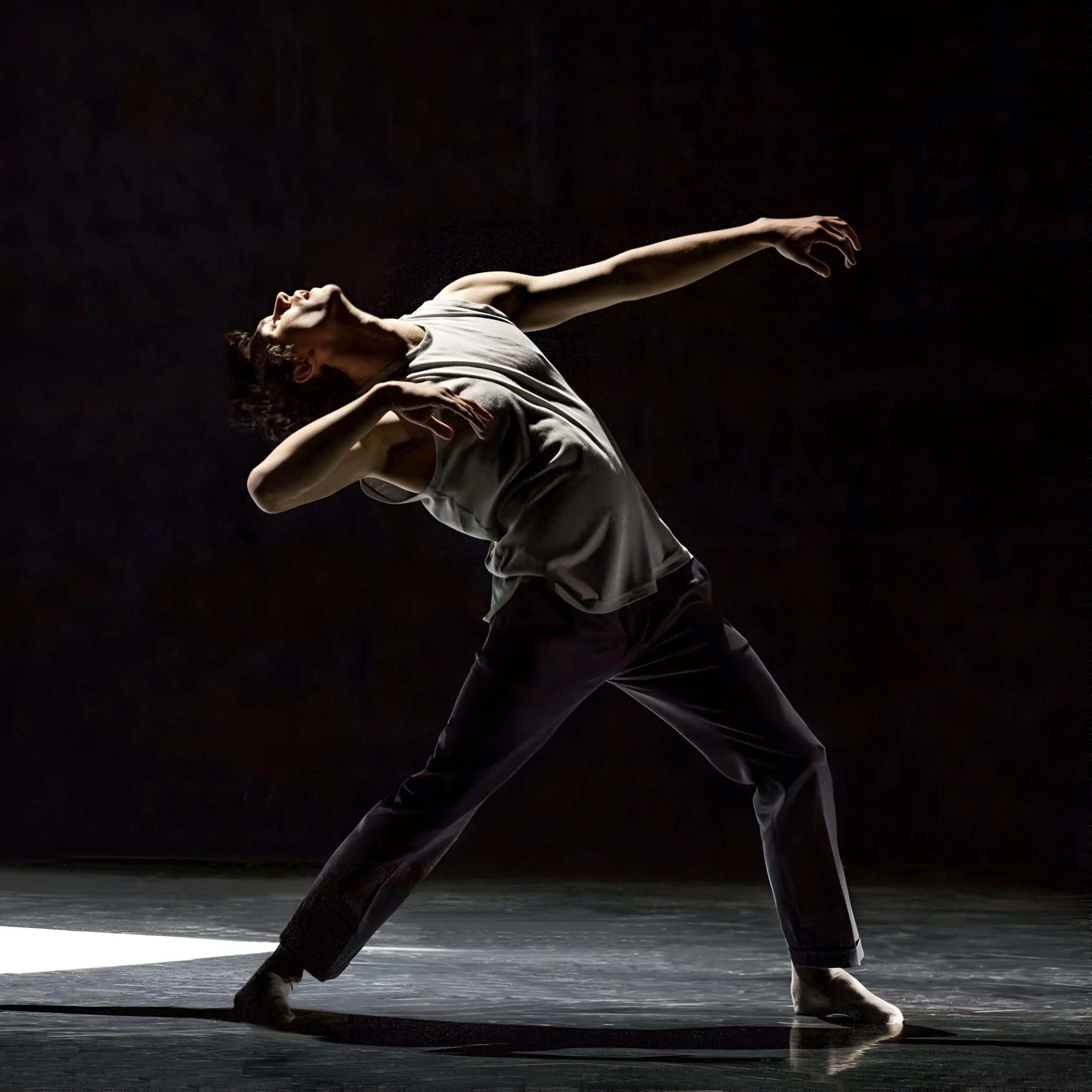 Une danseuse en mouvement dans une pièce sombre, portant une tenue grisâtre, avec un éclairage dramatique soulignant sa posture artistique.