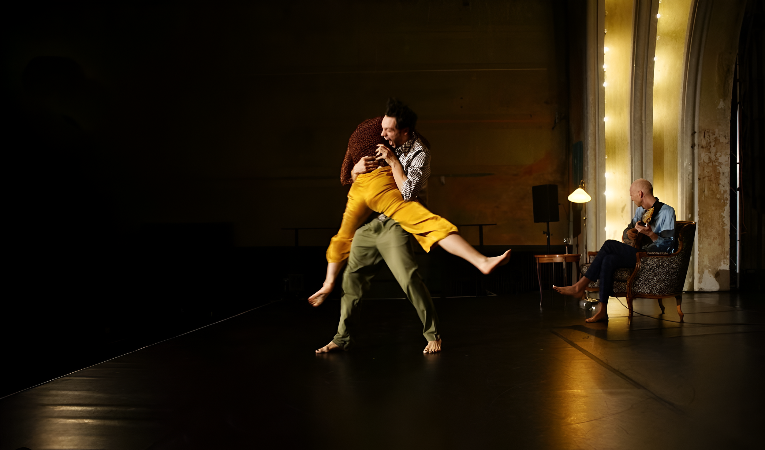 Deux danseurs en mouvement avec un musicien jouant de la guitare en arrière-plan, dans une pièce aux murs anciens et à l'éclairage chaud.