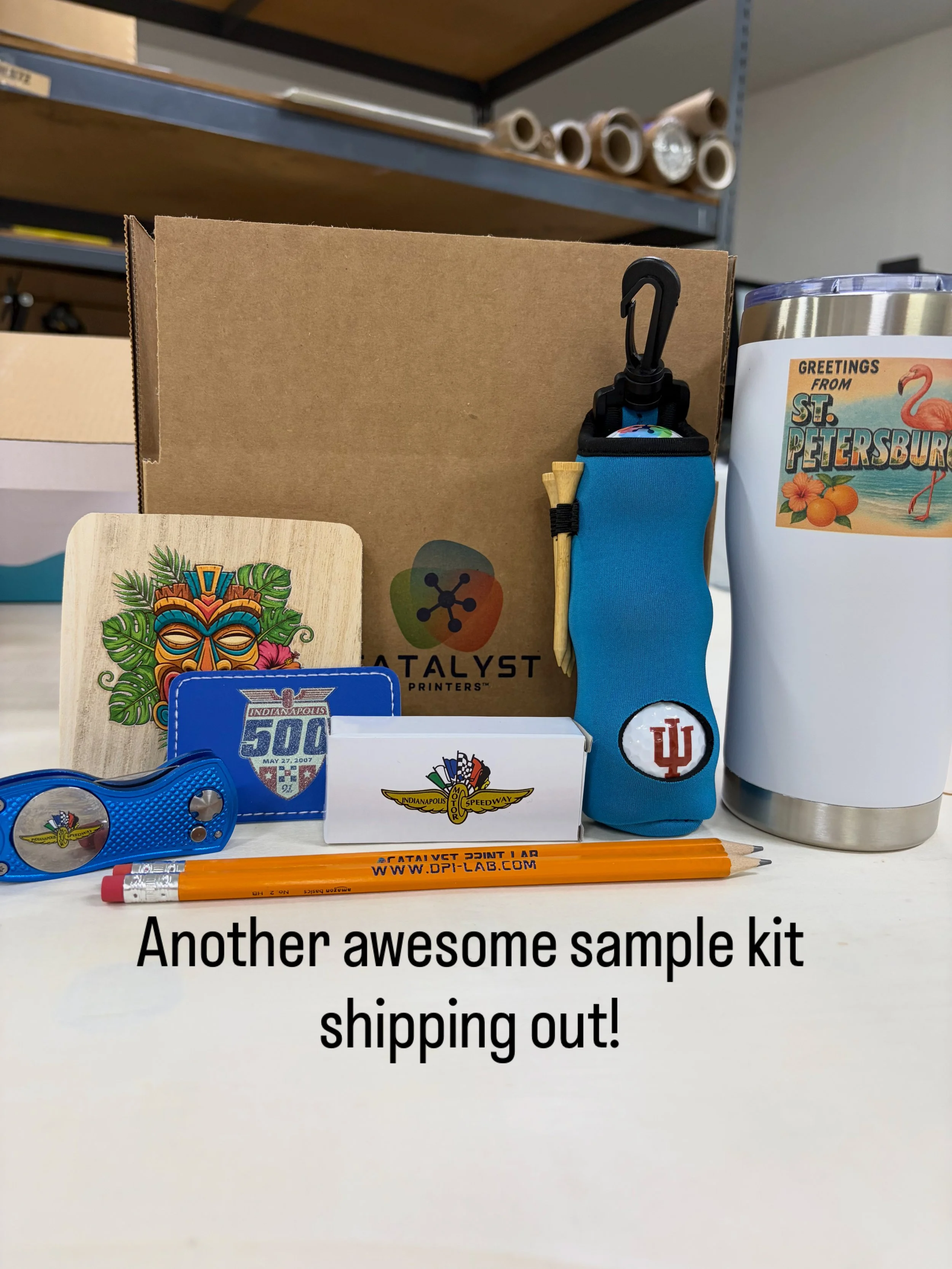 Sample kit with various items including a blue keychain, orange pencils, a souvenir card, a small box, a bottle opener, and a tumbler with a sticker, on a white surface in a storage room.