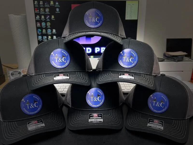 A pyramid of six black baseball caps with blue circular logos reading "T&C" on a desk in front of a computer monitor. Example of leather hat patches printed in full color with UV printer.