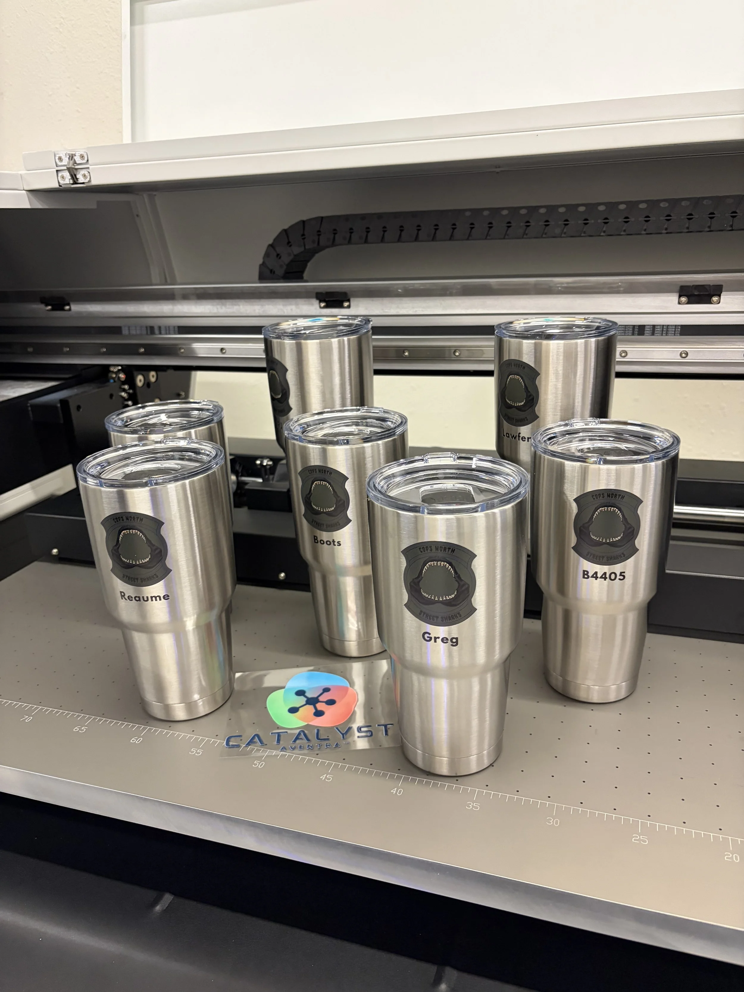 Seven stainless steel tumblers with customizable labels, placed on a work table in front of a laser engraving machine. Each tumbler has a black logo with text, showing different names and codes, with some labels reading 'Reauame', 'Boots', 'Greg', an