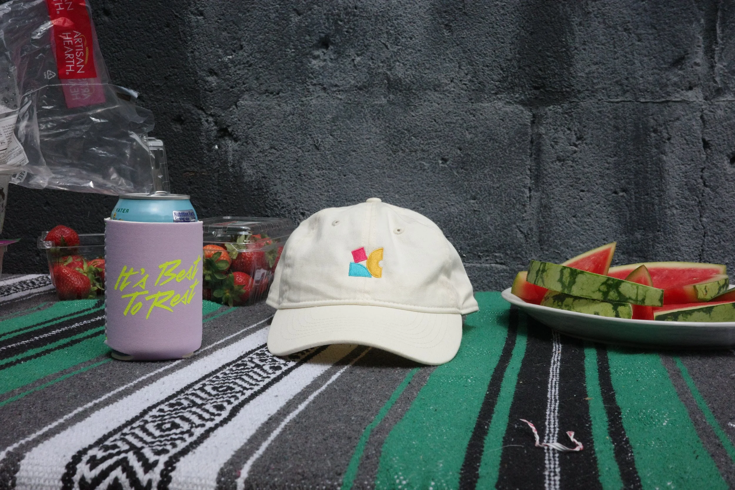 A white Chase Bliss Logo ball cap next to a "It's Best to Rest" Chase Bliss purple drink koozie.