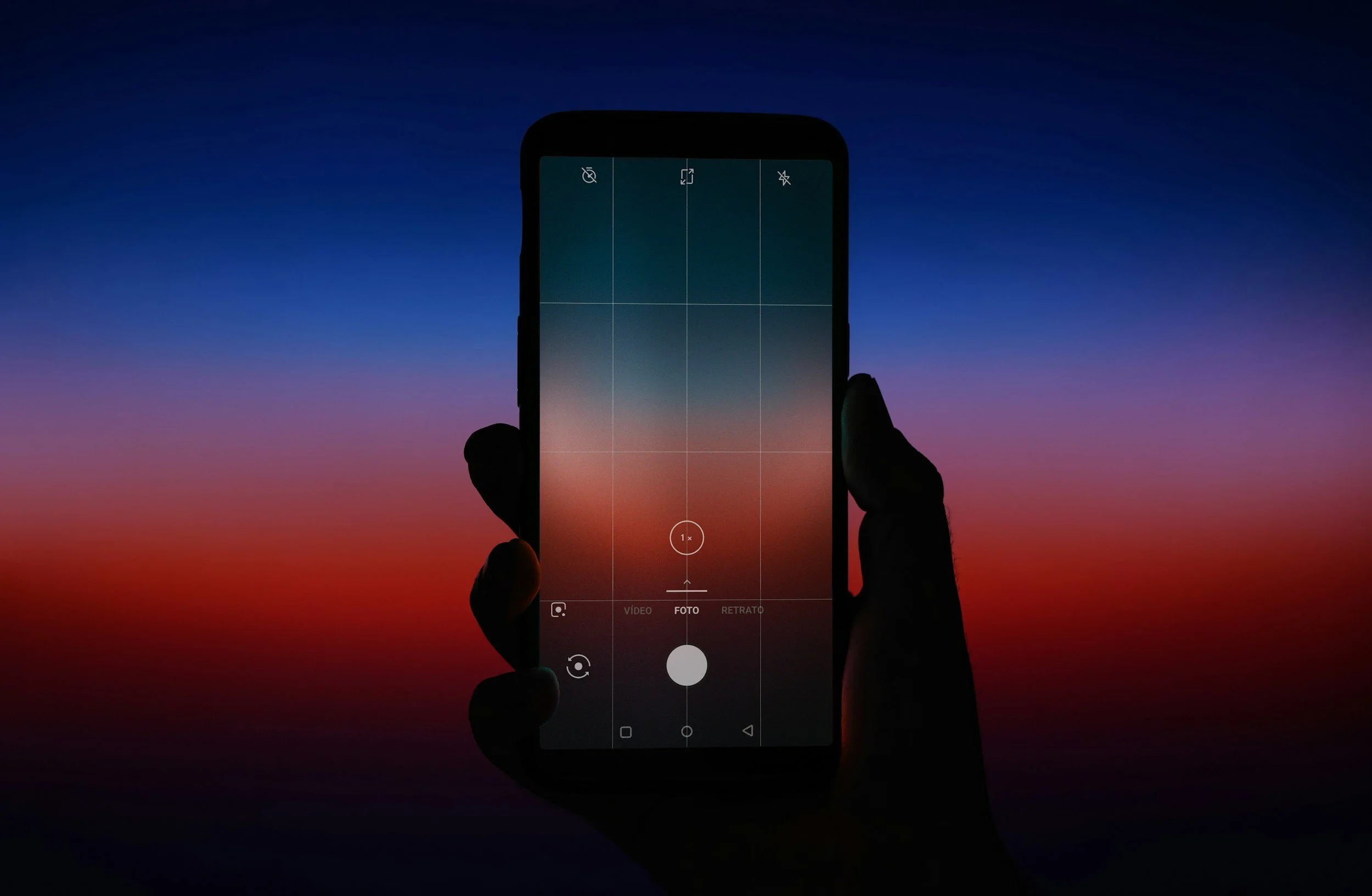 A person holding a smartphone taking a photo of a colorful sunset sky, with the phone's camera interface visible on the screen.