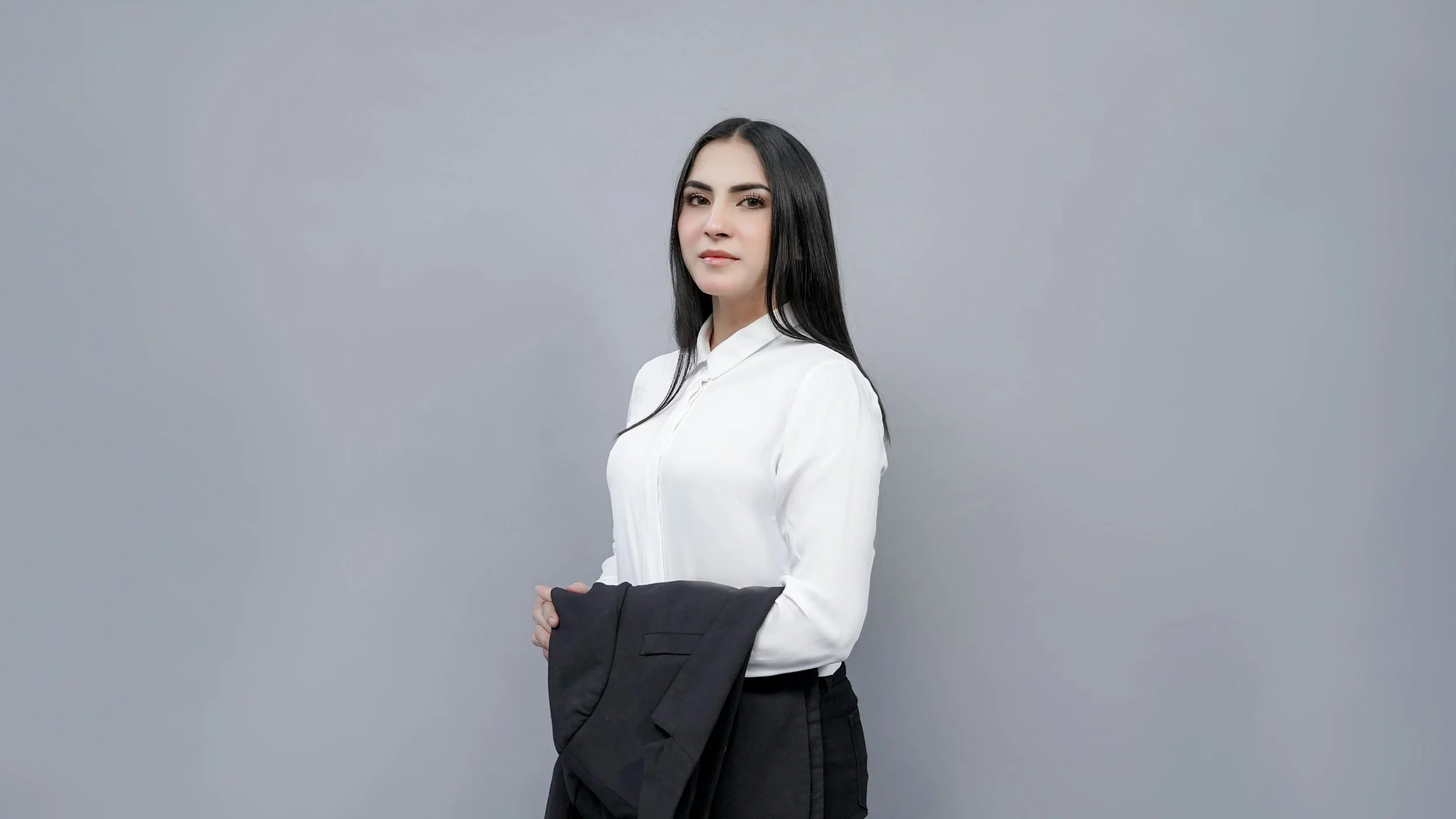 A young woman with long black hair wearing a white blouse holding a black blazer against a plain gray background.