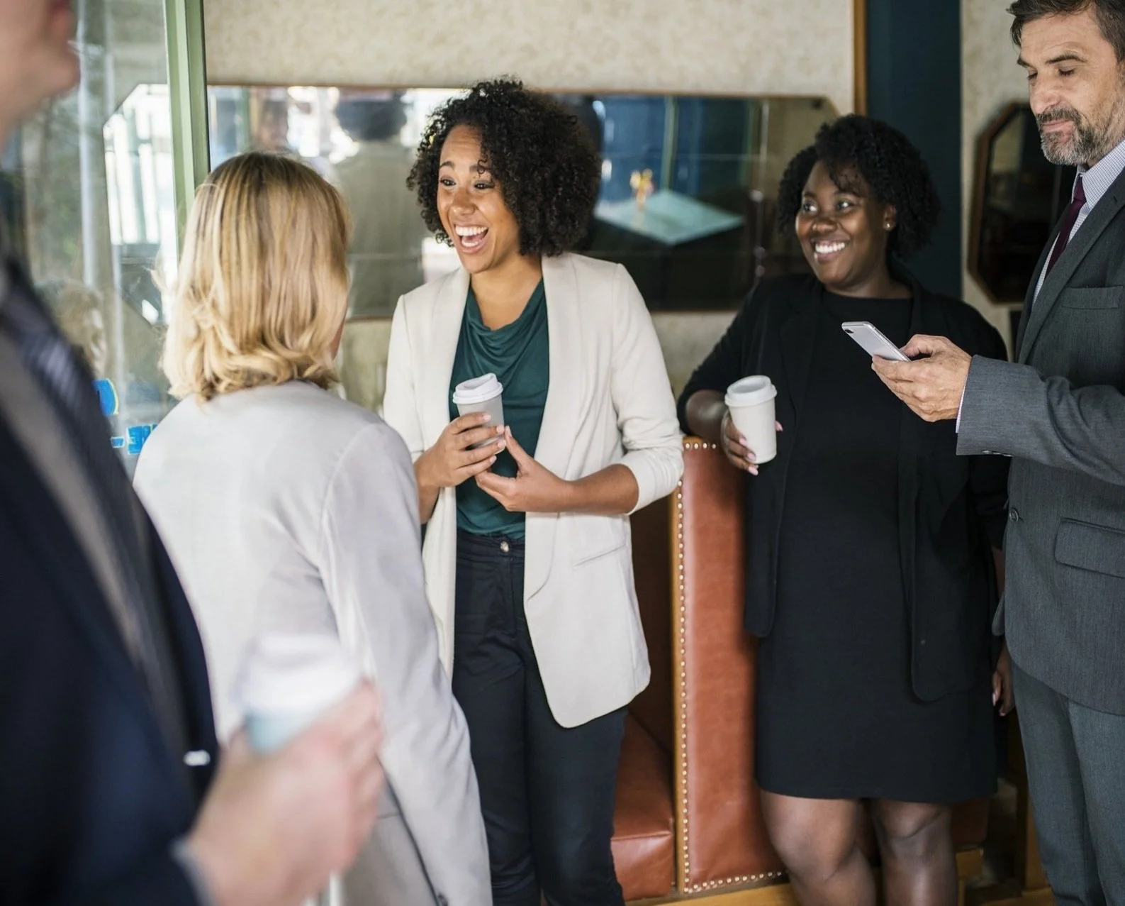 Group of five professionally dressed people socializing and smiling, holding coffee cups, inside a coffee shop or cafe.
