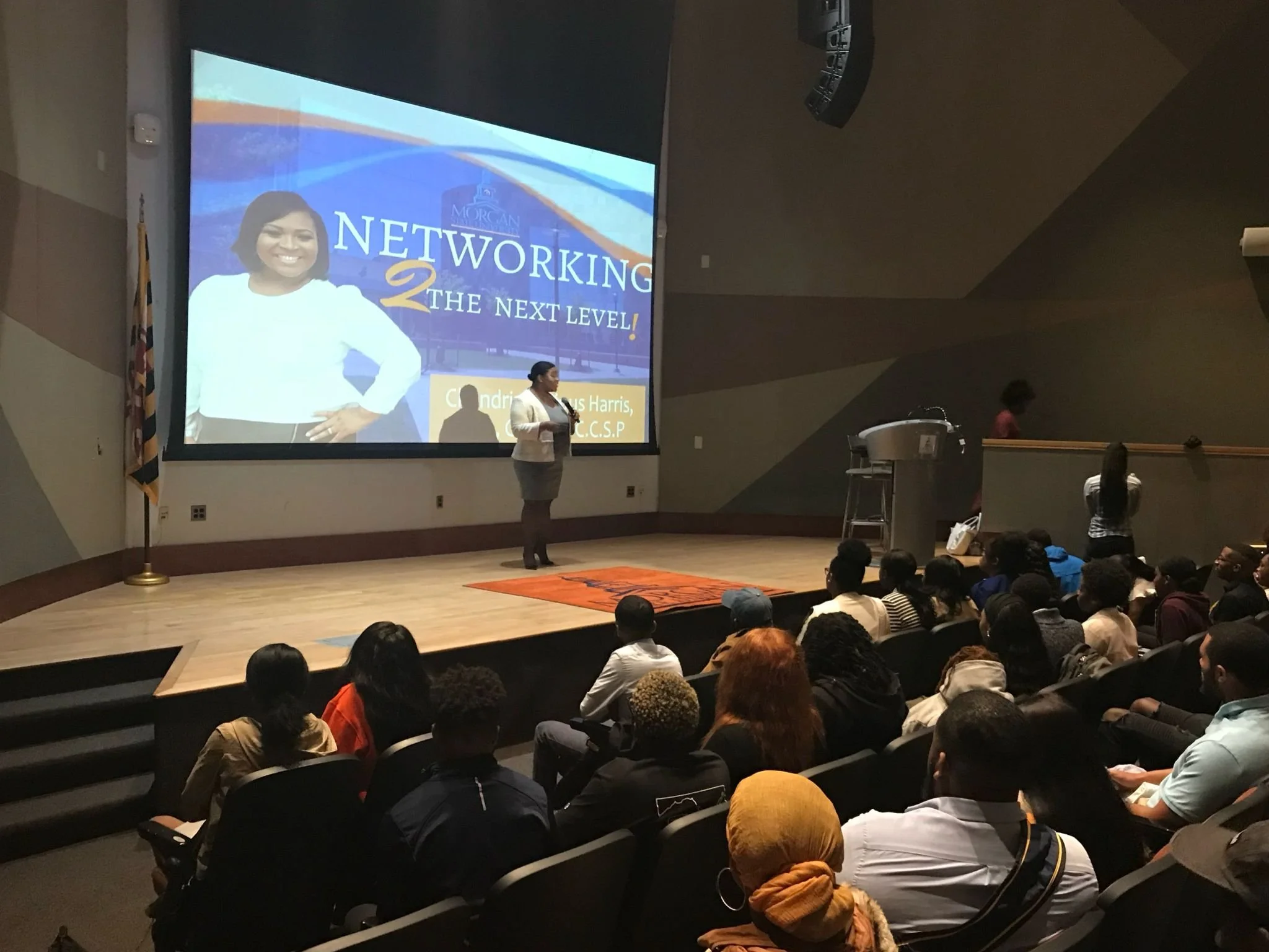 A woman giving a presentation on stage in front of a large screen that reads: 'Networking 2 the Next Level!' with an audience seated in a theater style setting.