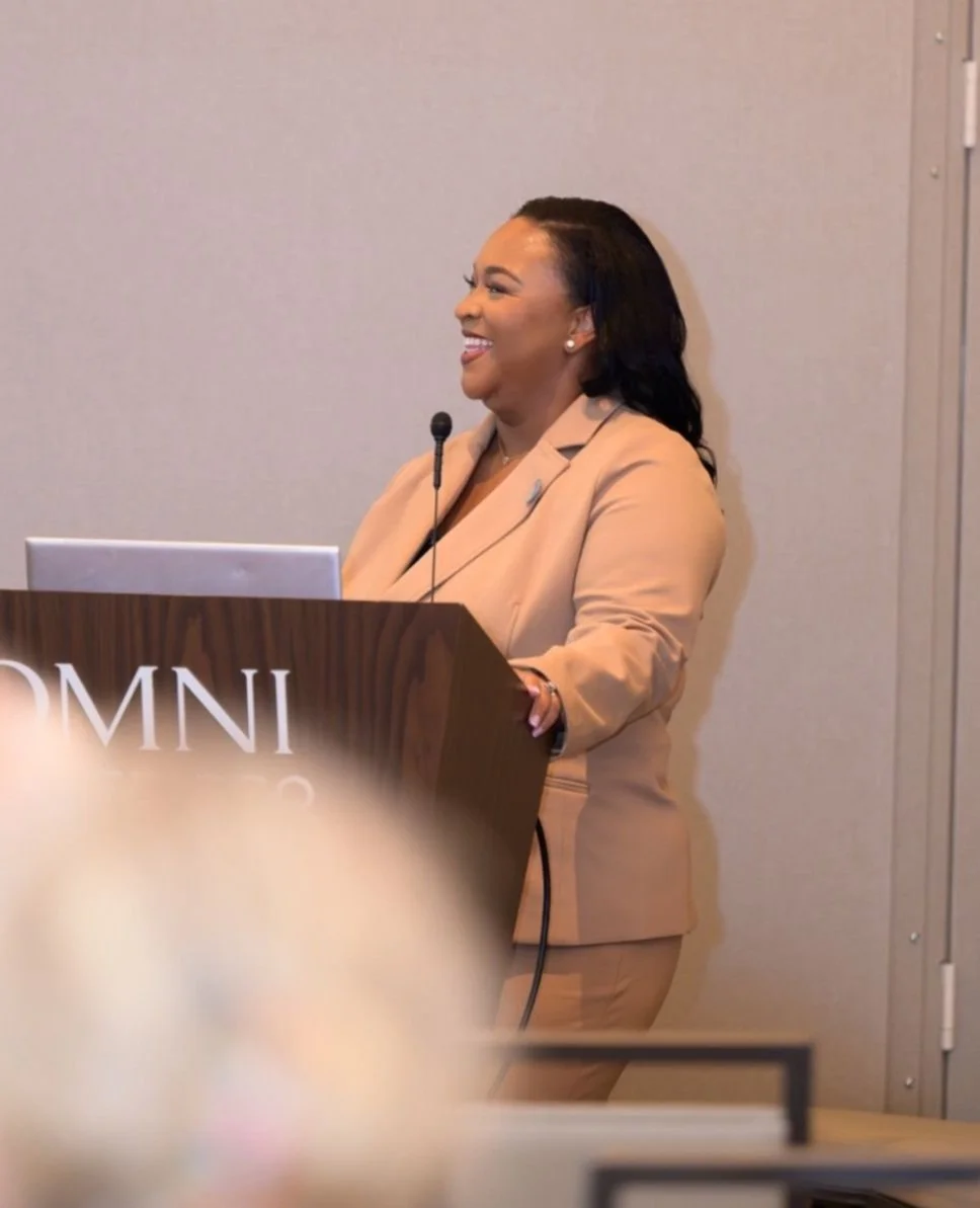 Woman giving a presentation at a conference, standing behind a wooden podium with a microphone, smiling, with the letters 'OMNI' visible on the front of the podium.