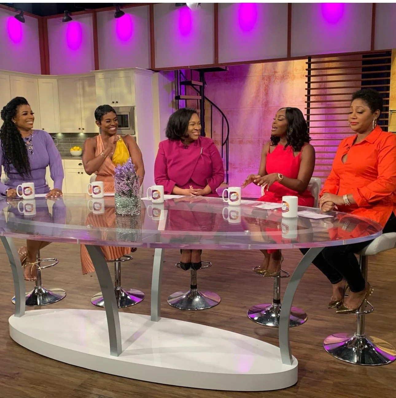 Five women sitting at a round glass table on a talk show set with purple, pink, and yellow lighting, kitchen background, engaged in conversation.