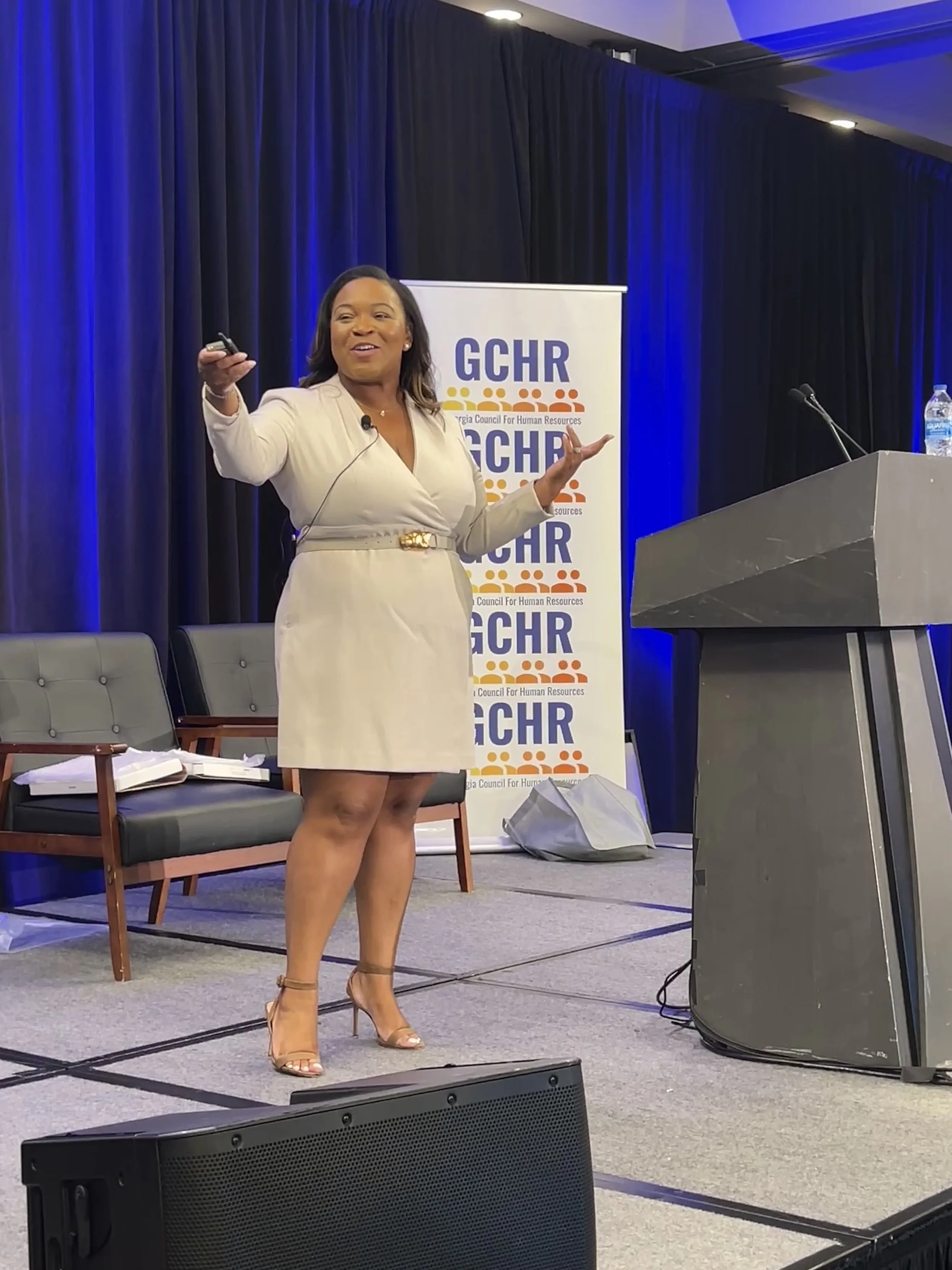 A woman in a beige dress and heels is speaking on stage with a microphone in her hand, standing in front of a GCHR banner at a conference.