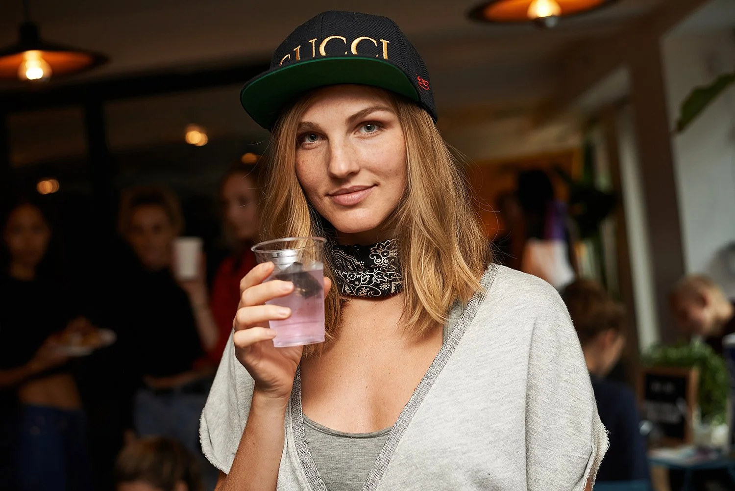 A young woman wearing a black Gucci cap, a bandana around her neck, and a light gray shirt, holding a plastic cup with a pink drink. She is smiling at the camera in a lively indoor setting with other people in the background.