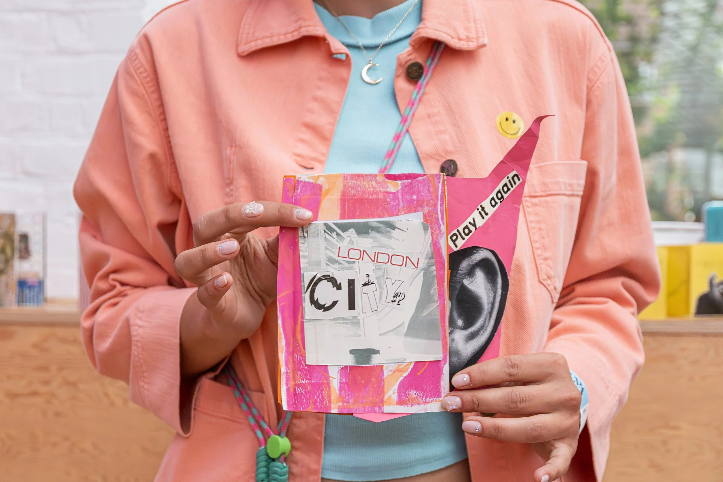 Person wearing a pink jacket, holding a colorful art piece with collage elements, including the word 'LONDON', a sticker saying 'Play it again', and various designs, in a setting with a blurred background.