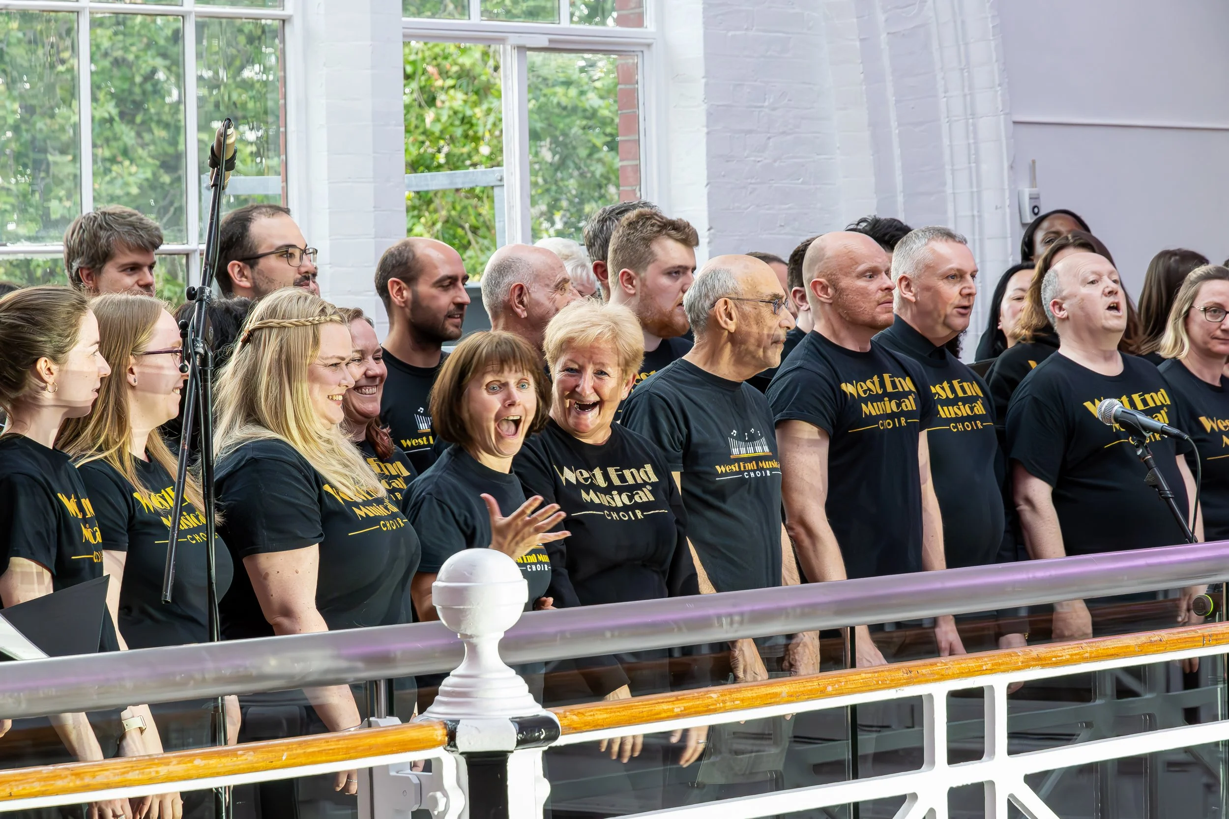 A choir group wearing black t-shirts with yellow text that says 'West End Musical Choir' singing during a performance or rehearsal indoors near a large window with greenery outside.