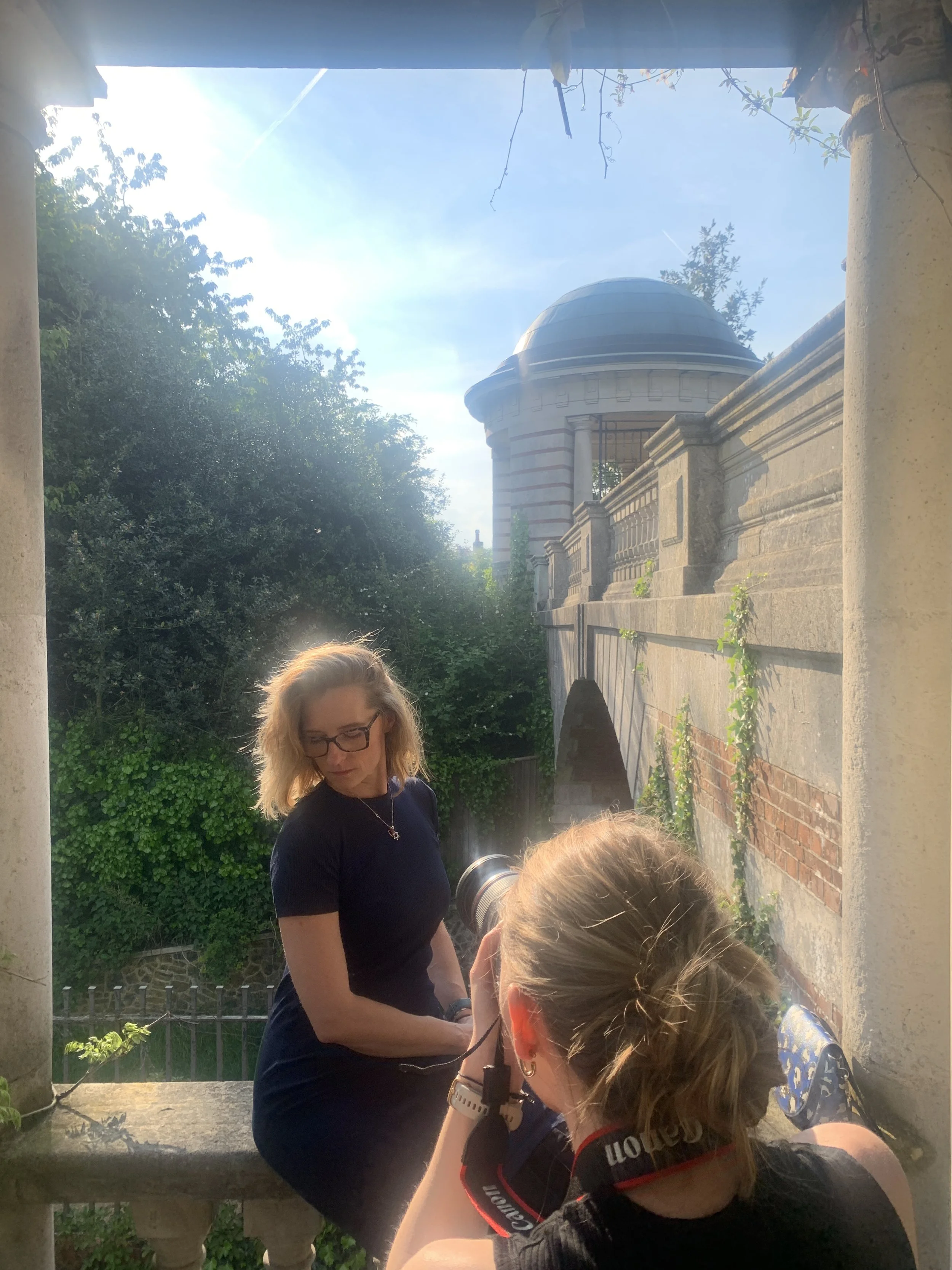 A woman posing for a photograph taken by another woman with a camera during a sunny day outside a historical building with a domed tower and stone walls surrounded by greenery.