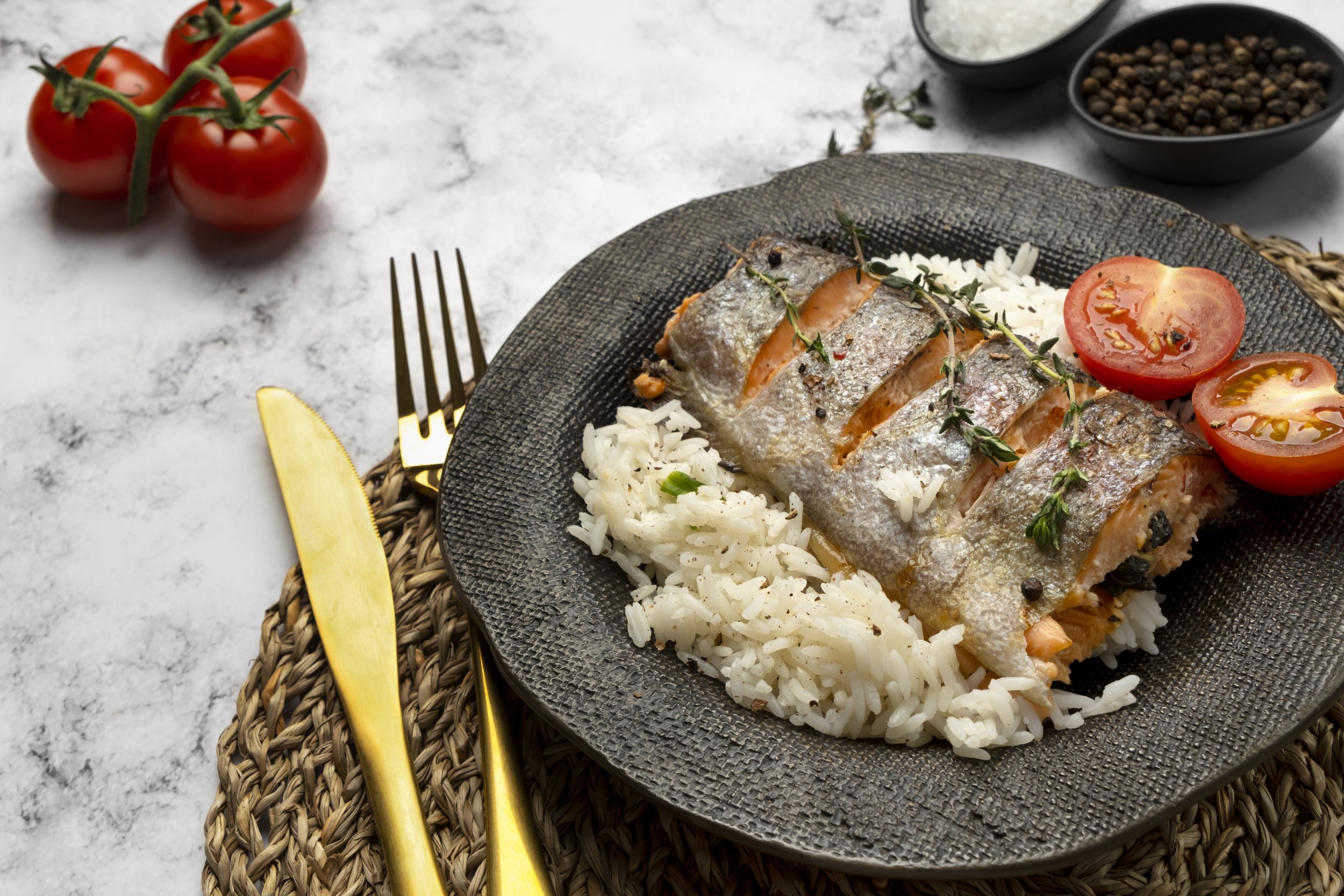 Cooked fish fillet on a bed of white rice, garnished with cherry tomatoes and fresh thyme, served on a black plate with gold utensils, on a gray marble surface.