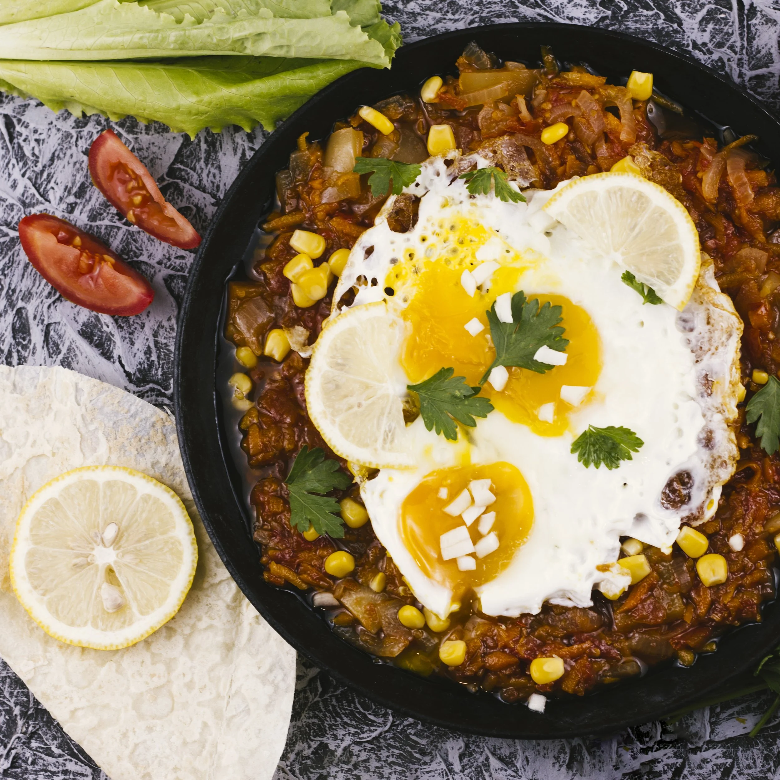 Cast-iron skillet with chili topped with two fried eggs, lemon slices, and cilantro, with fresh tomato slices, lettuce, lemon half, and tortilla chips on the side.
