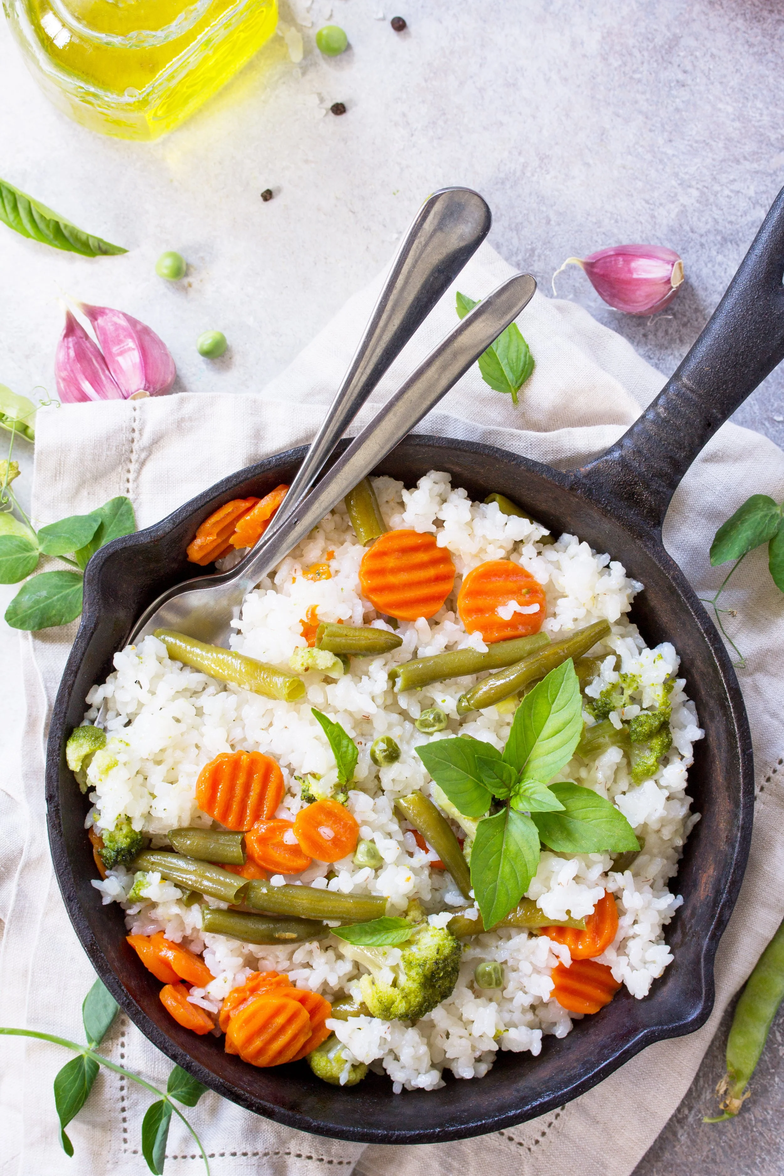 A cast iron skillet filled with cooked rice, green beans, carrots, and broccoli, garnished with fresh basil leaves, on a white cloth. Garlic cloves and a bottle of olive oil are in the background, along with scattered peppercorns and green peas.