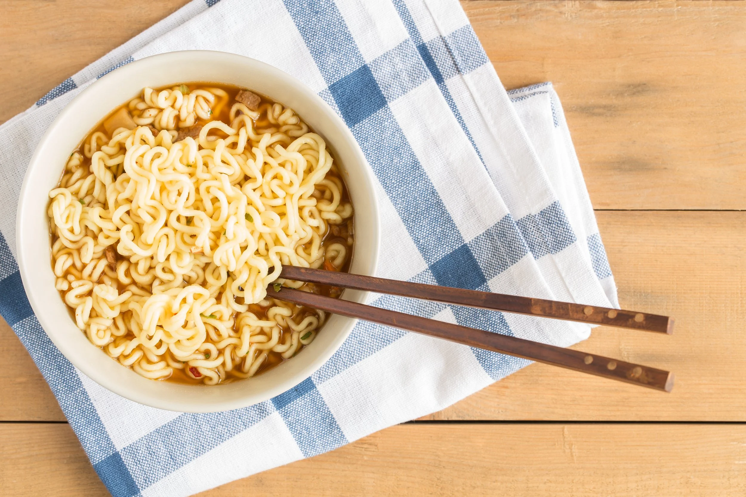 A bowl of cooked ramen noodles in broth with chopsticks resting on the edge, placed on a blue and white checkered cloth on a wooden surface.