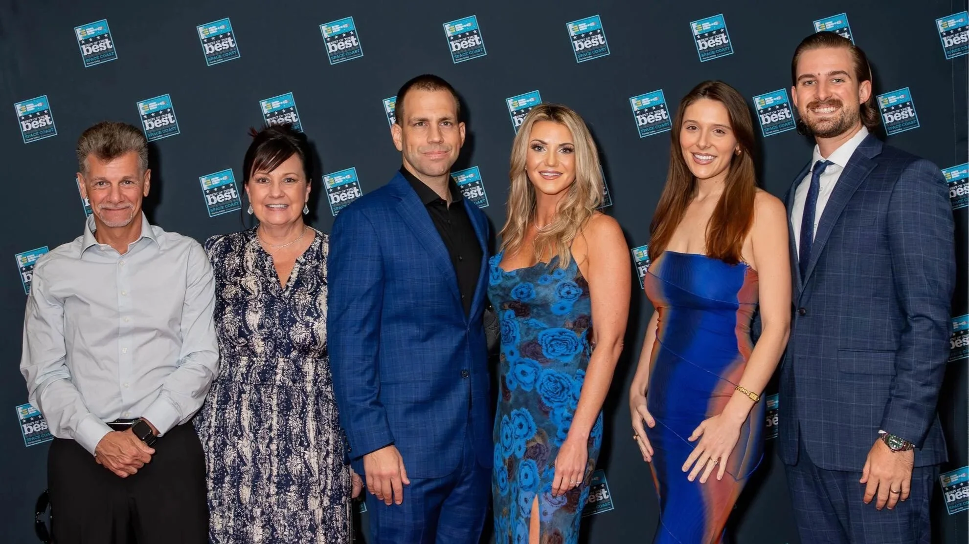 A group of six people—three men and three women—smile for a photo in front of a blue backdrop featuring "Best of the Best Space Coast" logos. They are dressed in semi-formal and formal attire.