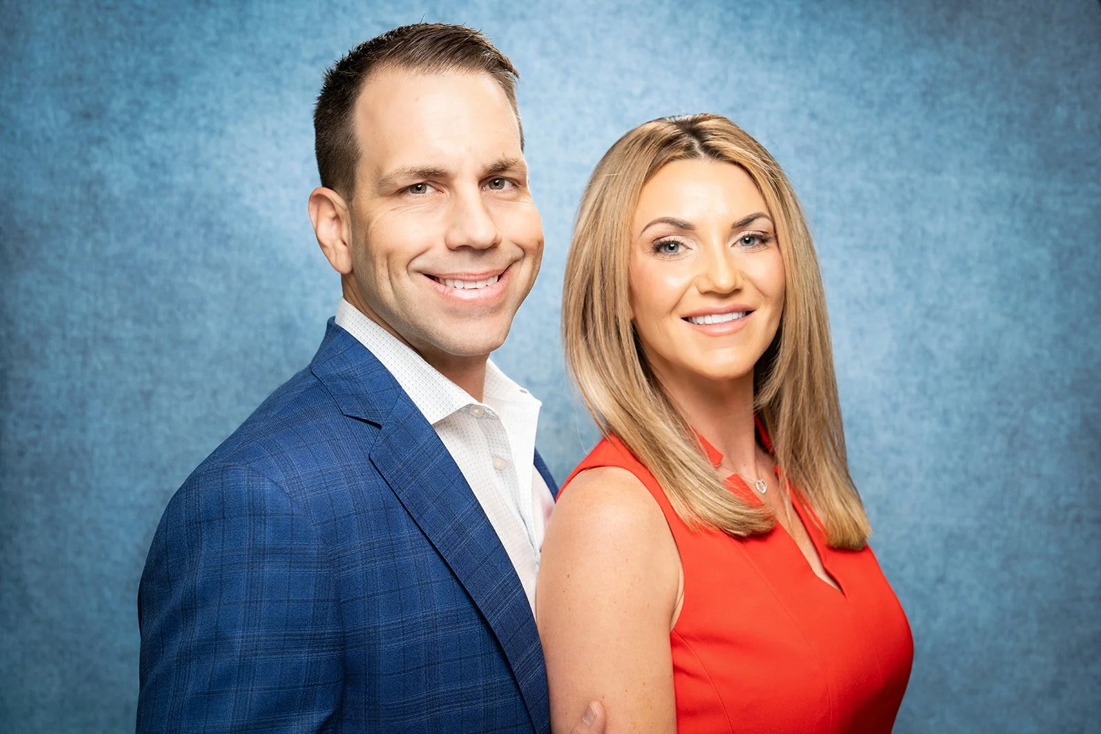 A man and a woman smiling, standing close together against a blue background. The man is wearing a blue suit jacket and white shirt. The woman is wearing a red sleeveless dress and has blonde hair.