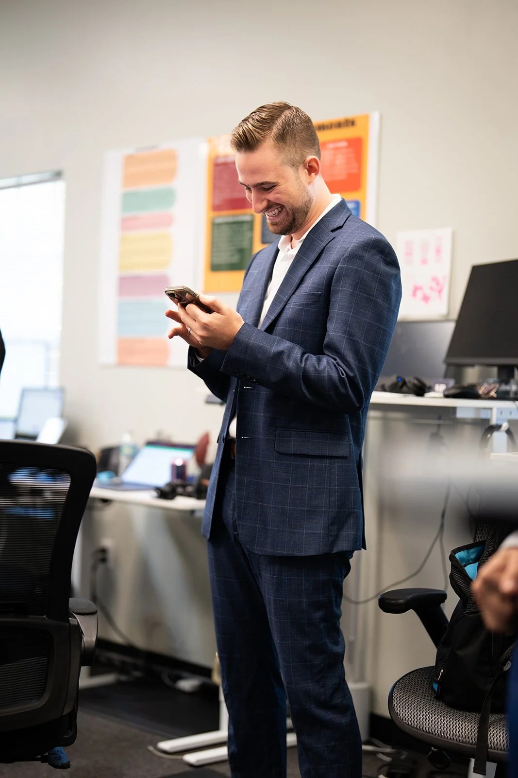 A man in a blue plaid suit is standing in an office, looking at his phone and smiling.
