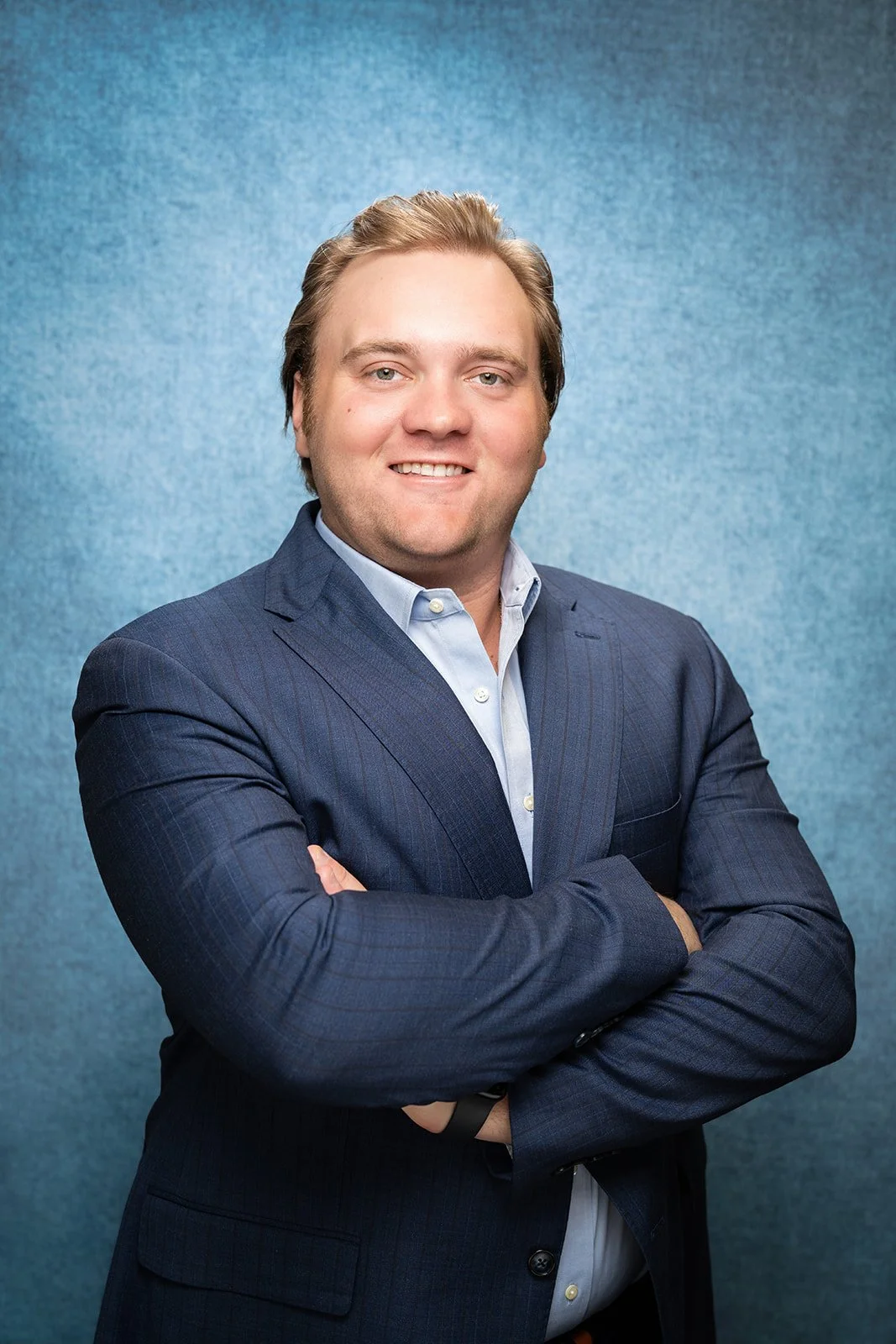 A smiling man with a beard and slicked-back hair, dressed in a gray business suit and white shirt, sitting with hands clasped in front of a blurred office background.