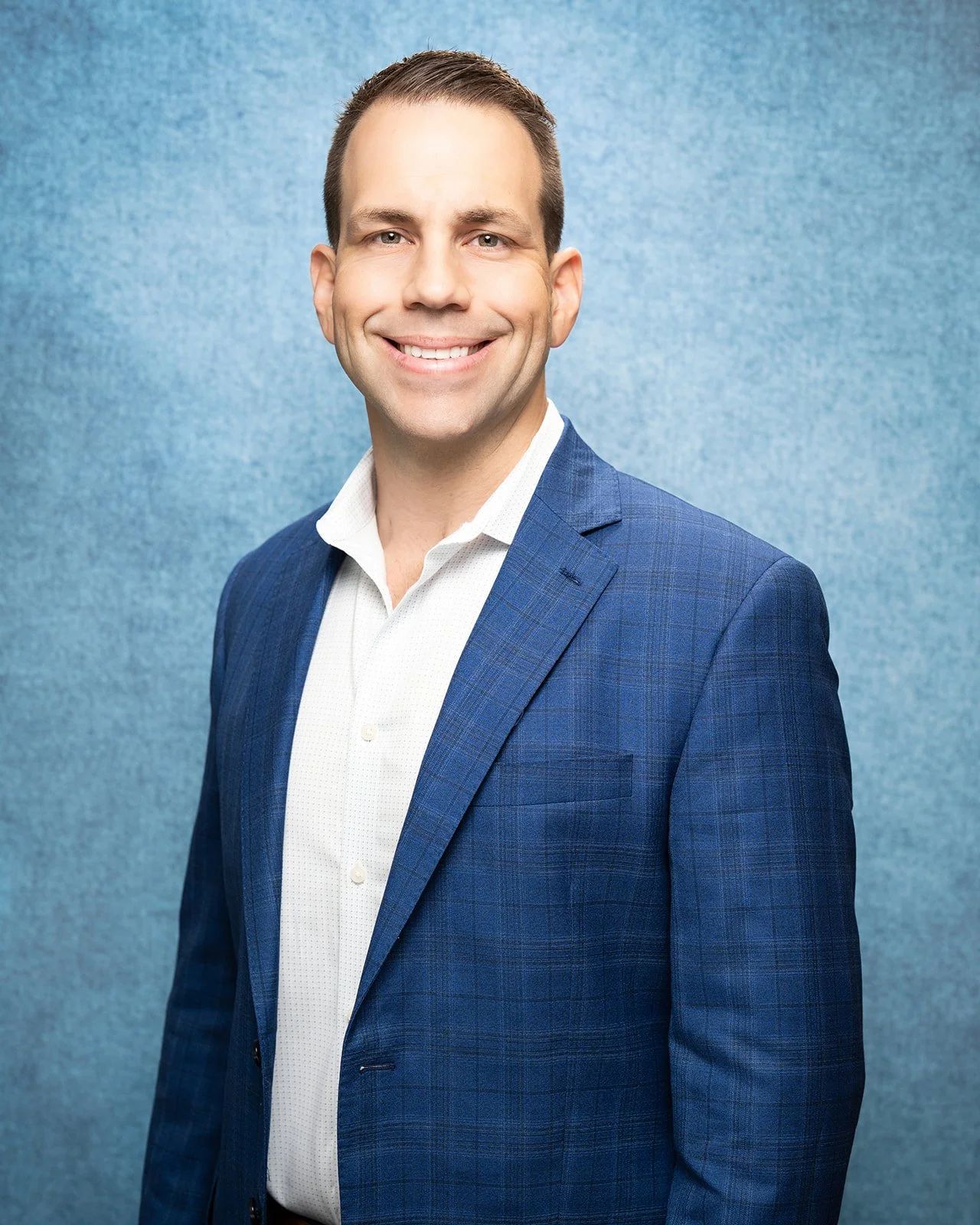 A man with short brown hair in a white shirt and blue suit jacket smiling against a blue textured background.