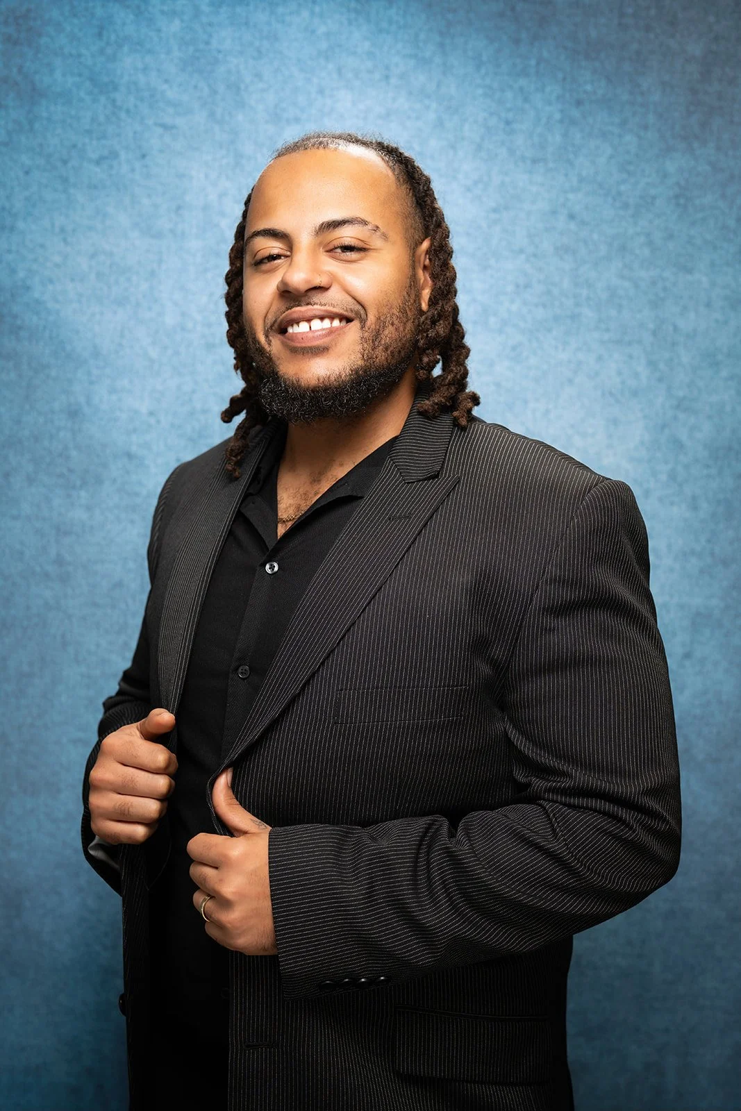 A smiling man wearing a black suit, burgundy shirt, and black patterned tie, standing against a plain white wall.