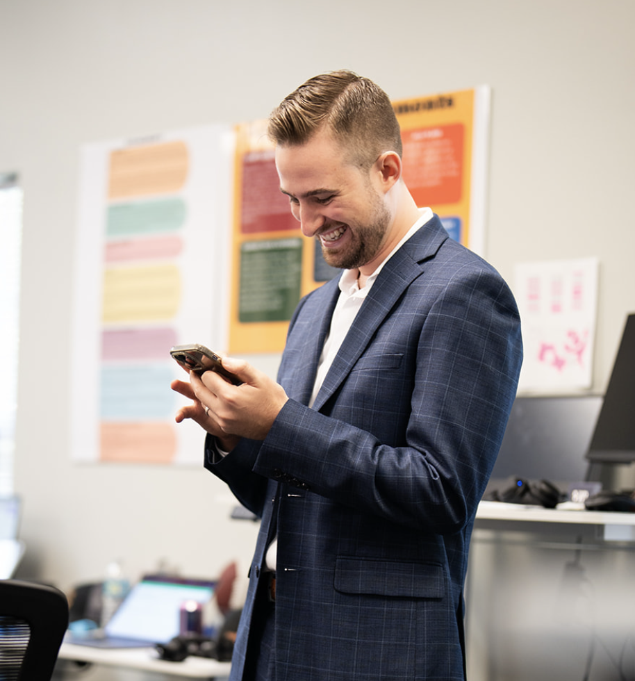 Smiling man in business suit looking at his phone in an office with colorful wall posters.