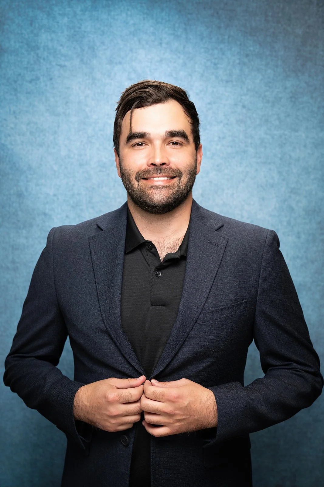 A young man with dark hair and a beard, wearing a black suit jacket and white shirt, standing against a blue background.