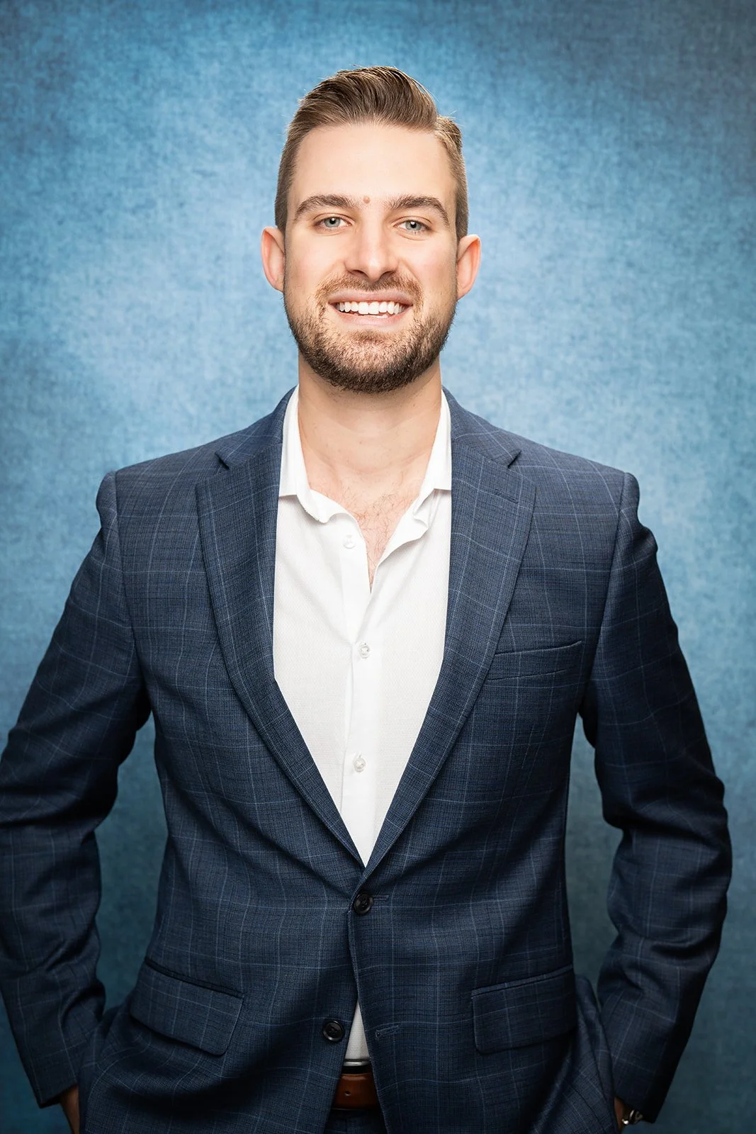 A young man with light brown hair and a beard, wearing a dark blue plaid suit jacket and a white dress shirt, smiling in front of a neutral background.