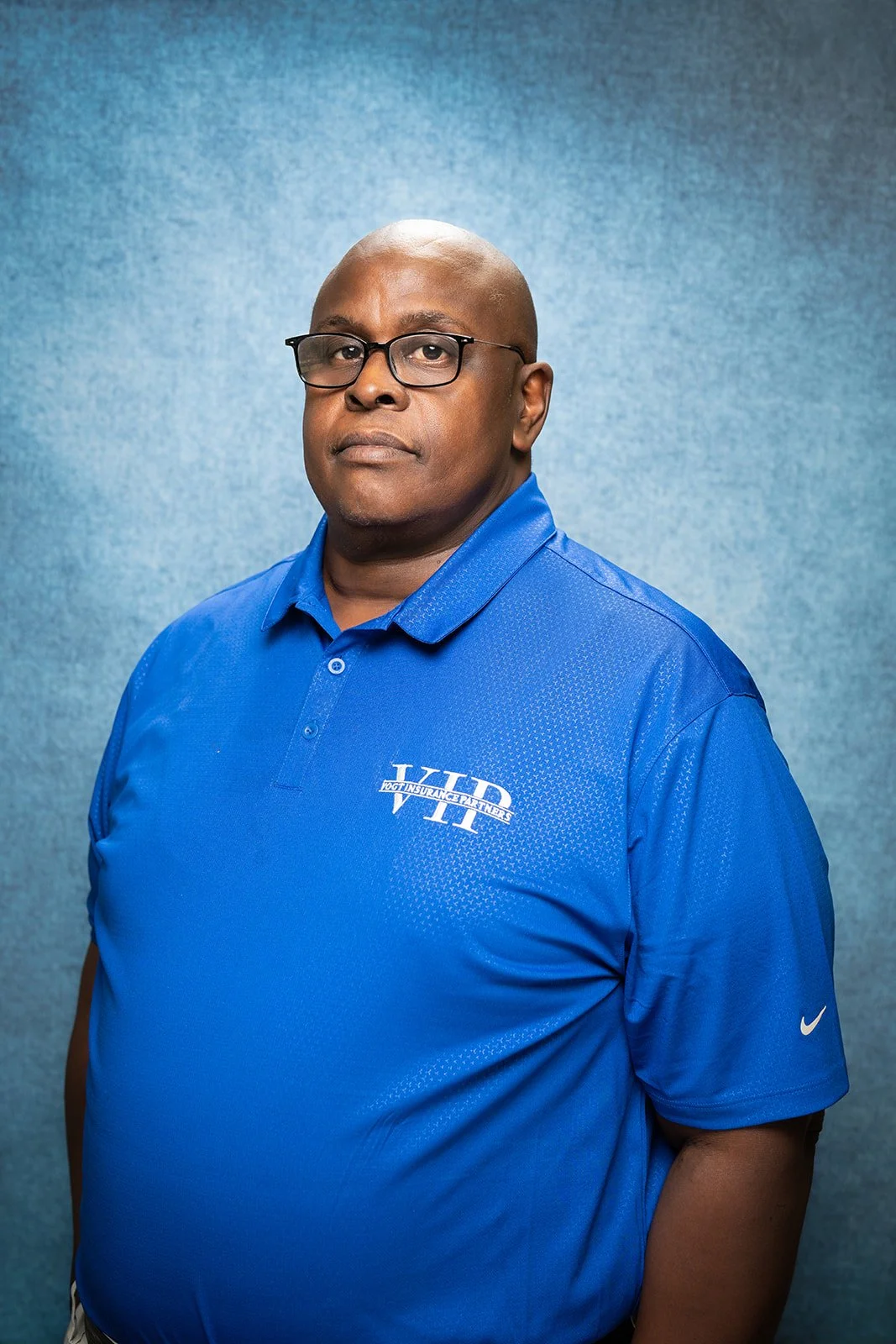 A man with glasses and a serious expression is wearing a blue polo shirt with a logo that reads 'LTD Dog Insurance Partners' and a small Nike logo on the sleeve, standing against a textured blue background.