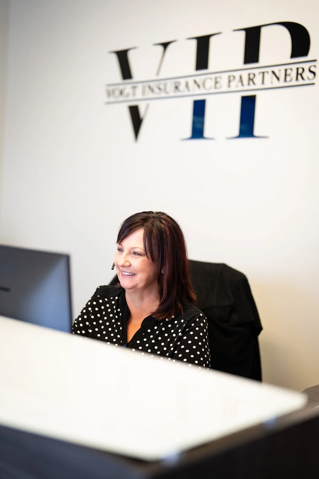 Woman with dark hair and polka dot blouse sitting at a desk, smiling at her computer in an office with the VLI logo on the wall behind her.