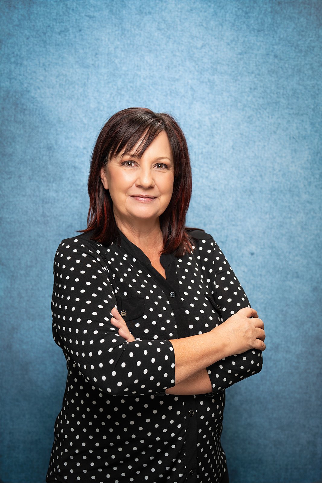 A smiling woman with shoulder-length dark brown hair and hoop earrings, wearing a patterned top, posed against a gray background.