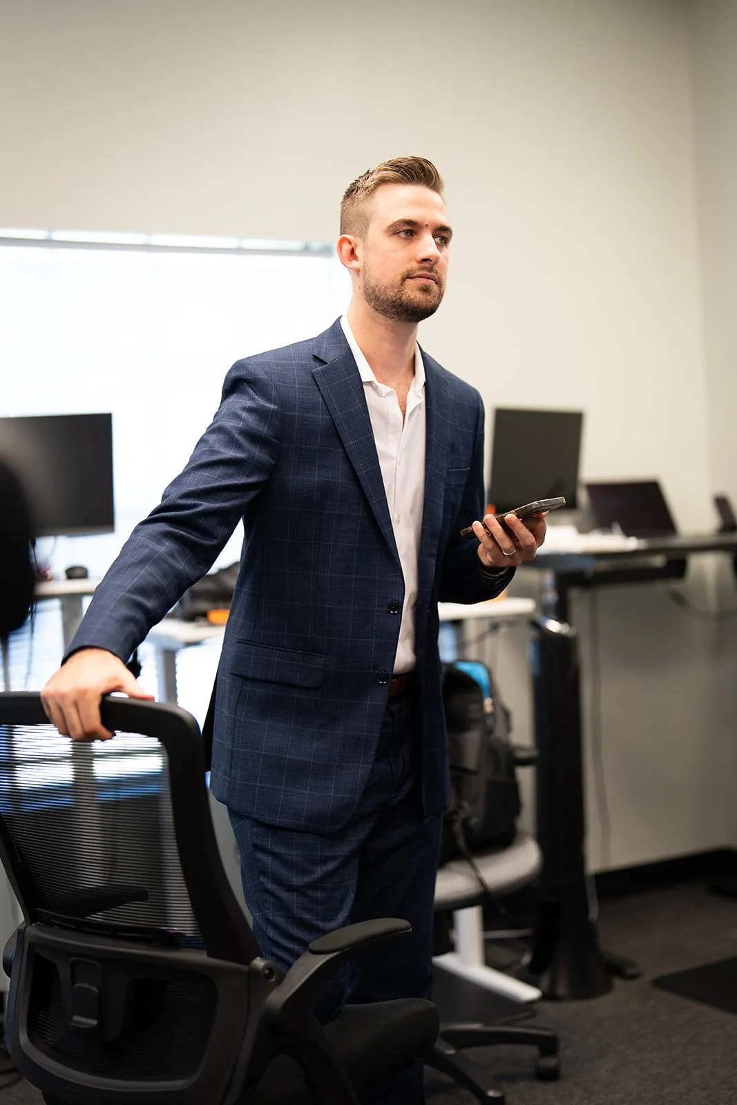 A man in a checkered navy blazer and white shirt standing in an office, holding a smartphone in his right hand, with office desks and computers in the background.
